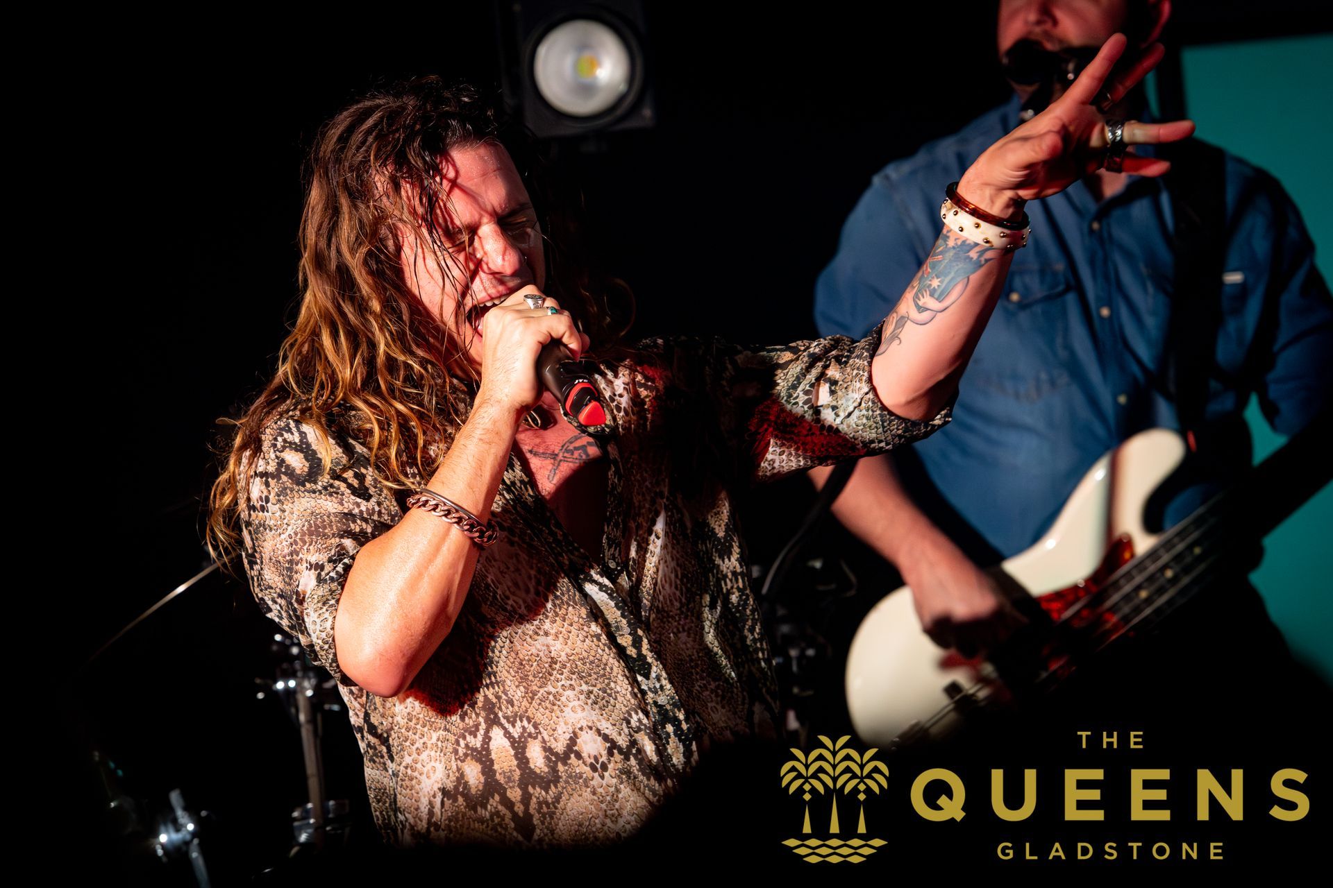 Singer performing onstage with a microphone beside a guitarist, with “The Queens” logo in the corner