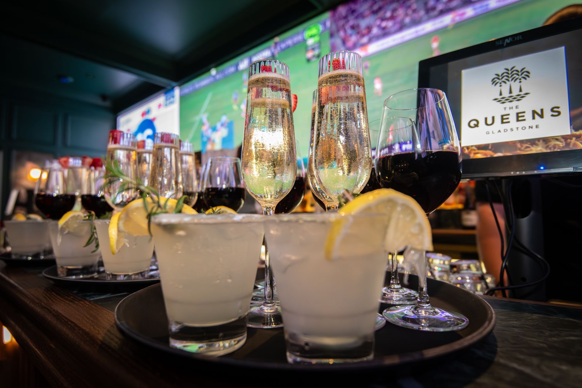 A tray of mixed drinks and wine glasses on a bar. A TV screen in the background.