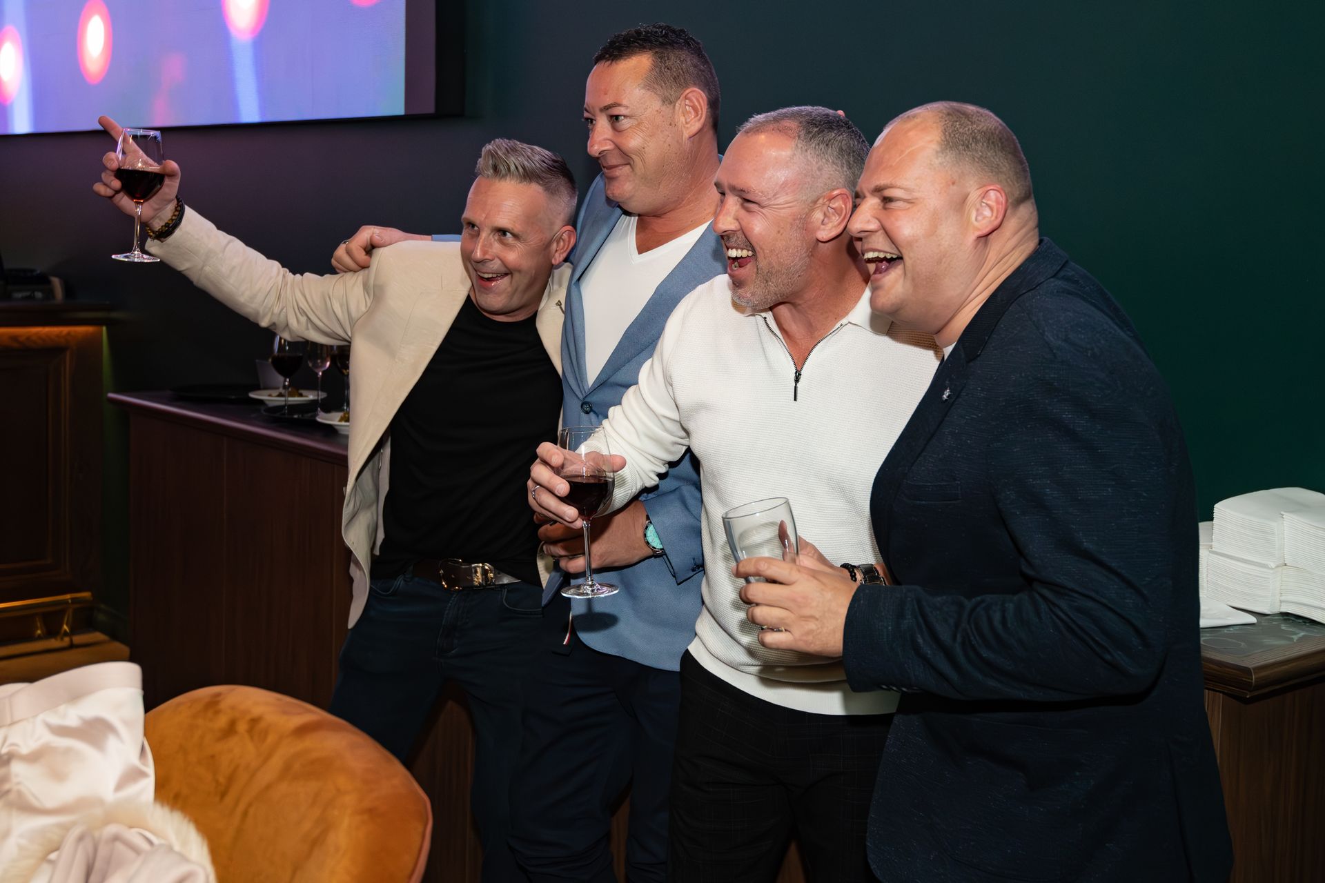 Four men at an event, posing and smiling. One raises a glass of red wine, another holds a drink.