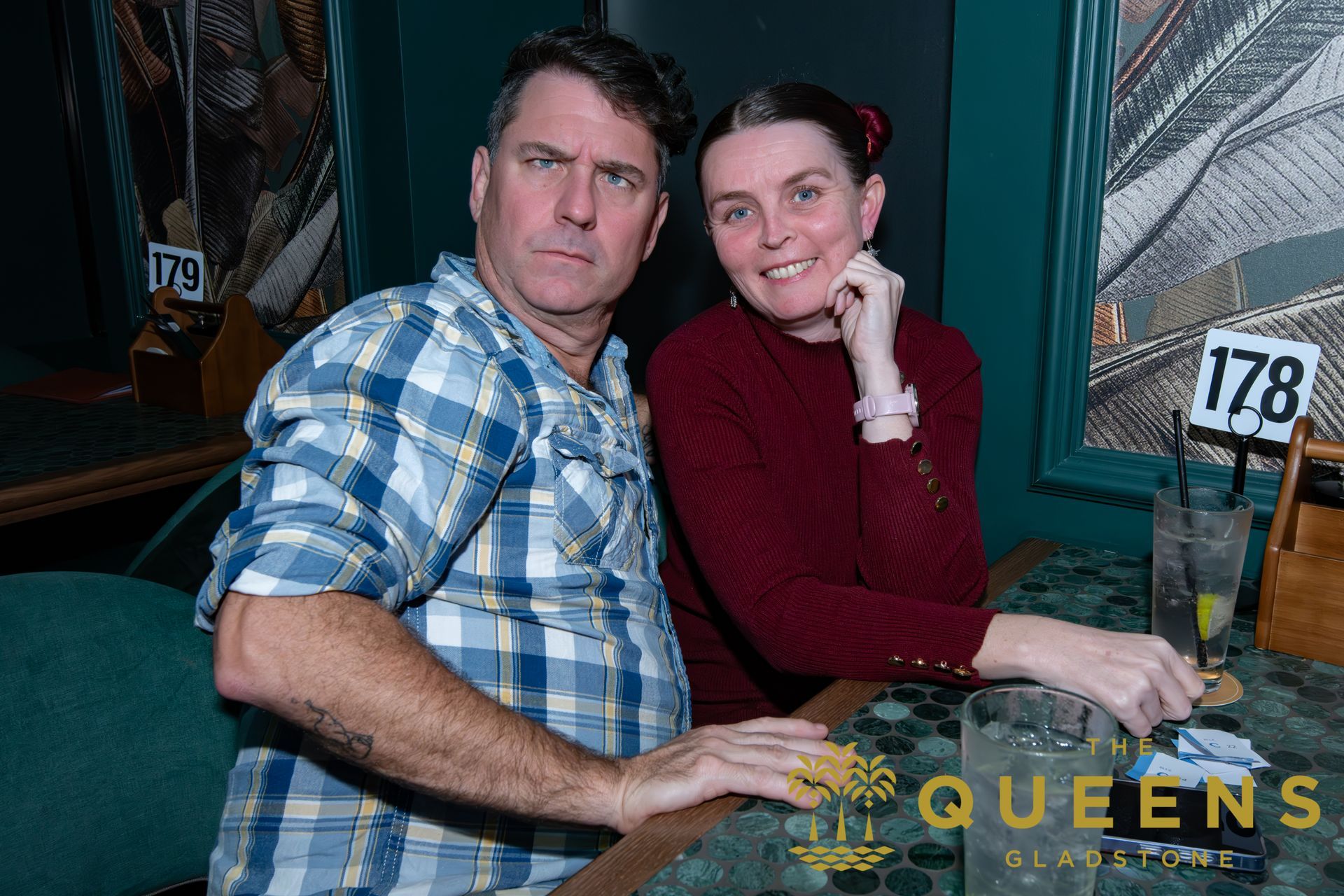 Two people seated together at a table, posing indoors with a QUEENS sign in the background.