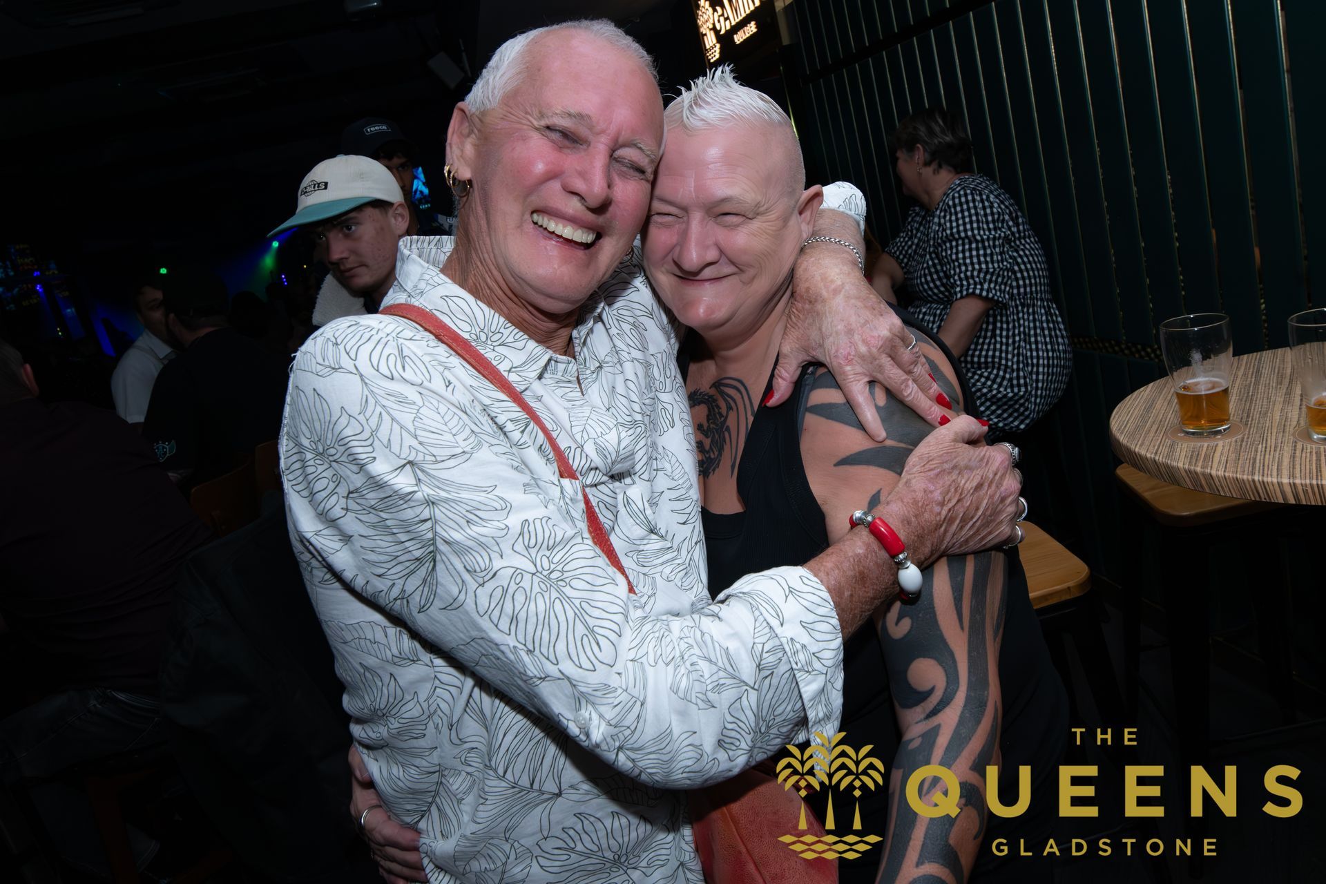 Two smiling men embracing at a dark nightclub table, with drinks and a “The Queens” sign visible.