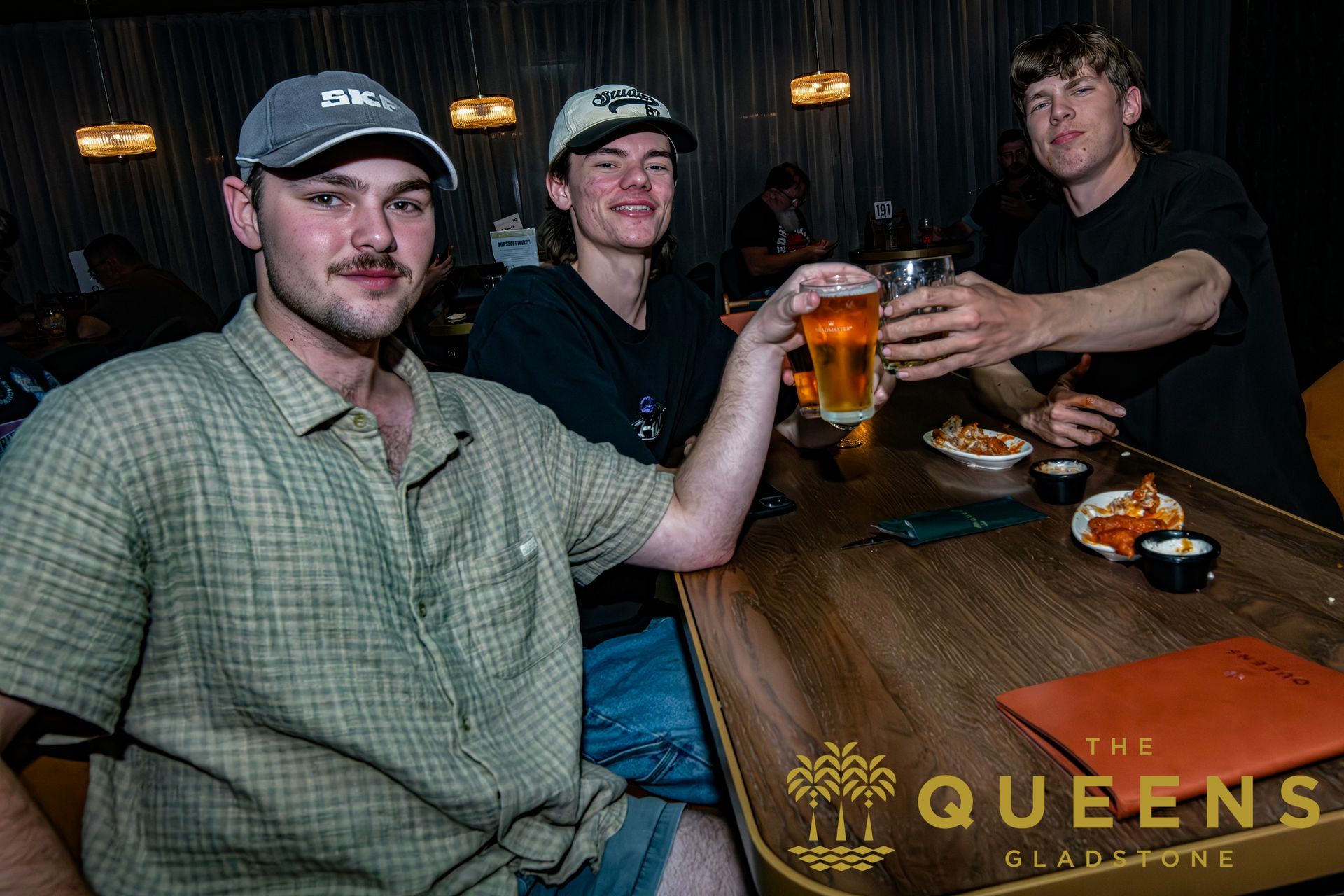 Three people toasting drinks at a bar table, with a “QUEENS” sign visible on the table front.