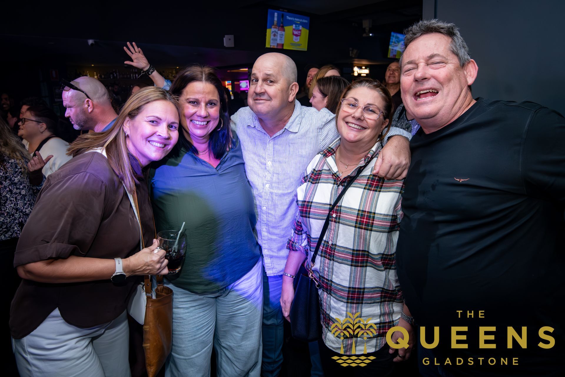 Five smiling friends posing together at The Queens Gladstone, with one woman waving behind them