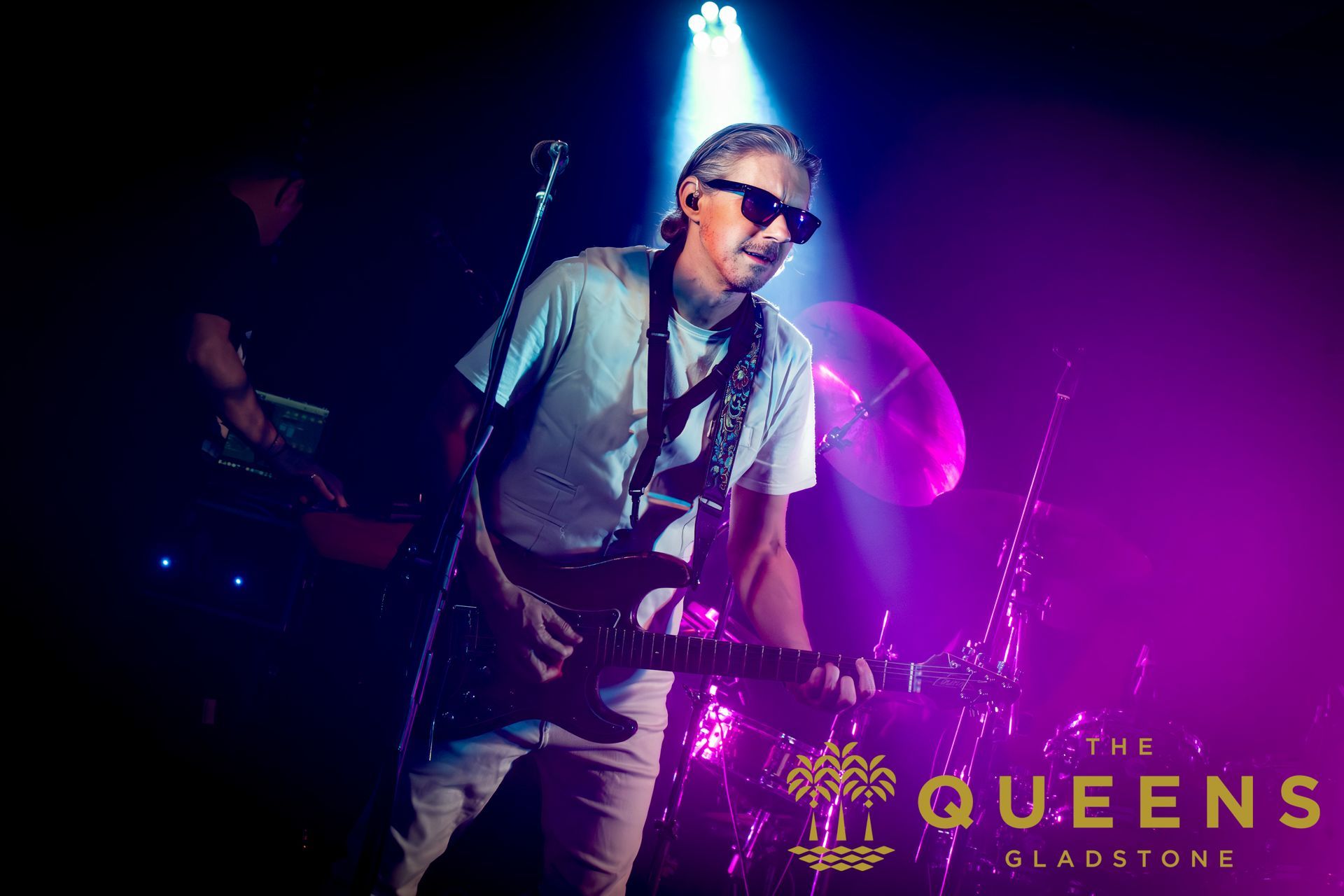 Singer performing on a dimly lit stage under blue and purple lights, with “QUEENS” signage visible.