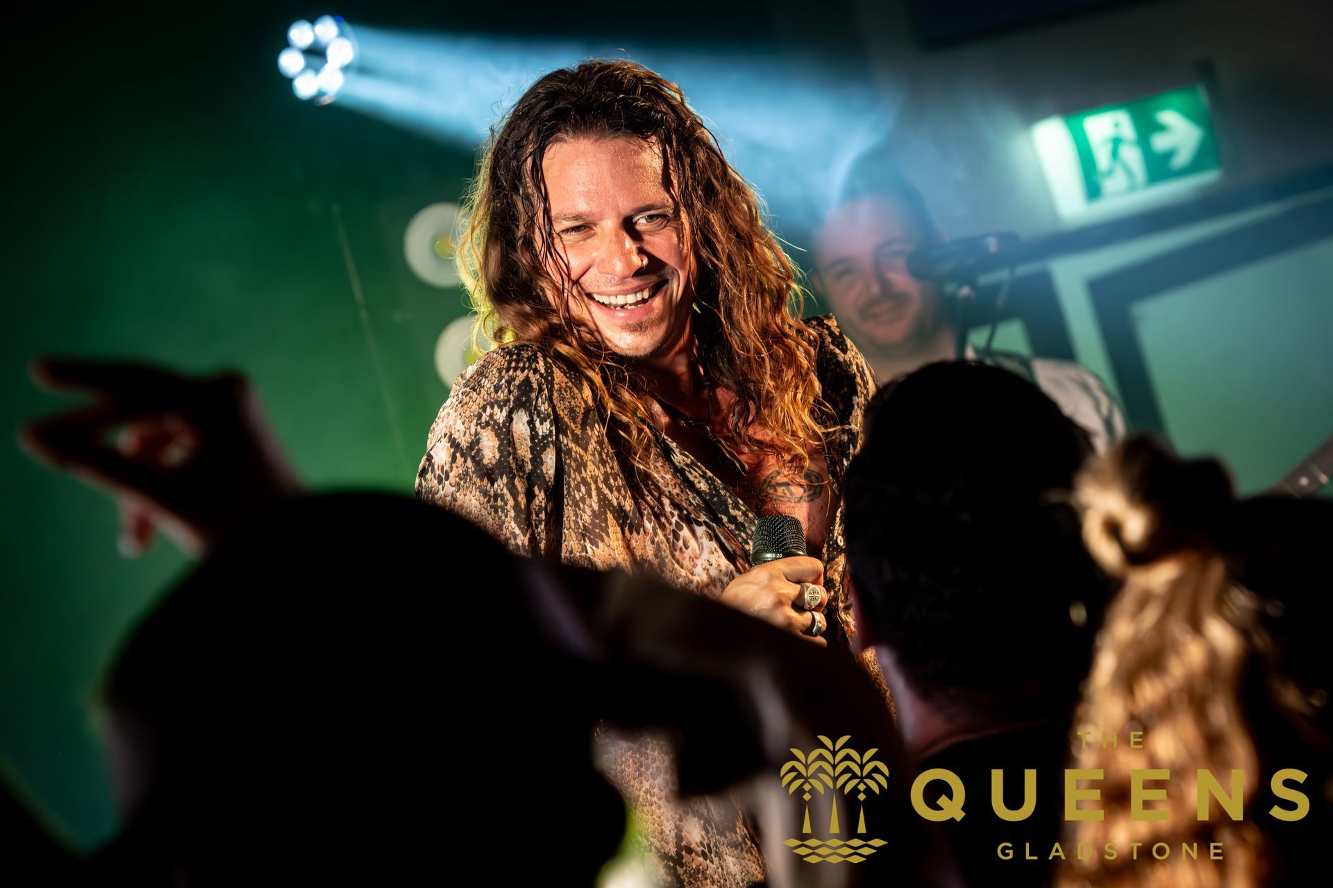 Smiling performer with long curly hair in a dim club, with crowd silhouettes and green exit sign in background