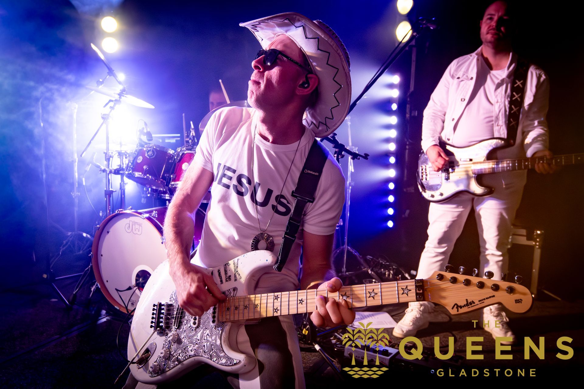 Rock band performing on stage, singer in white hat and shirt playing guitar under purple lights