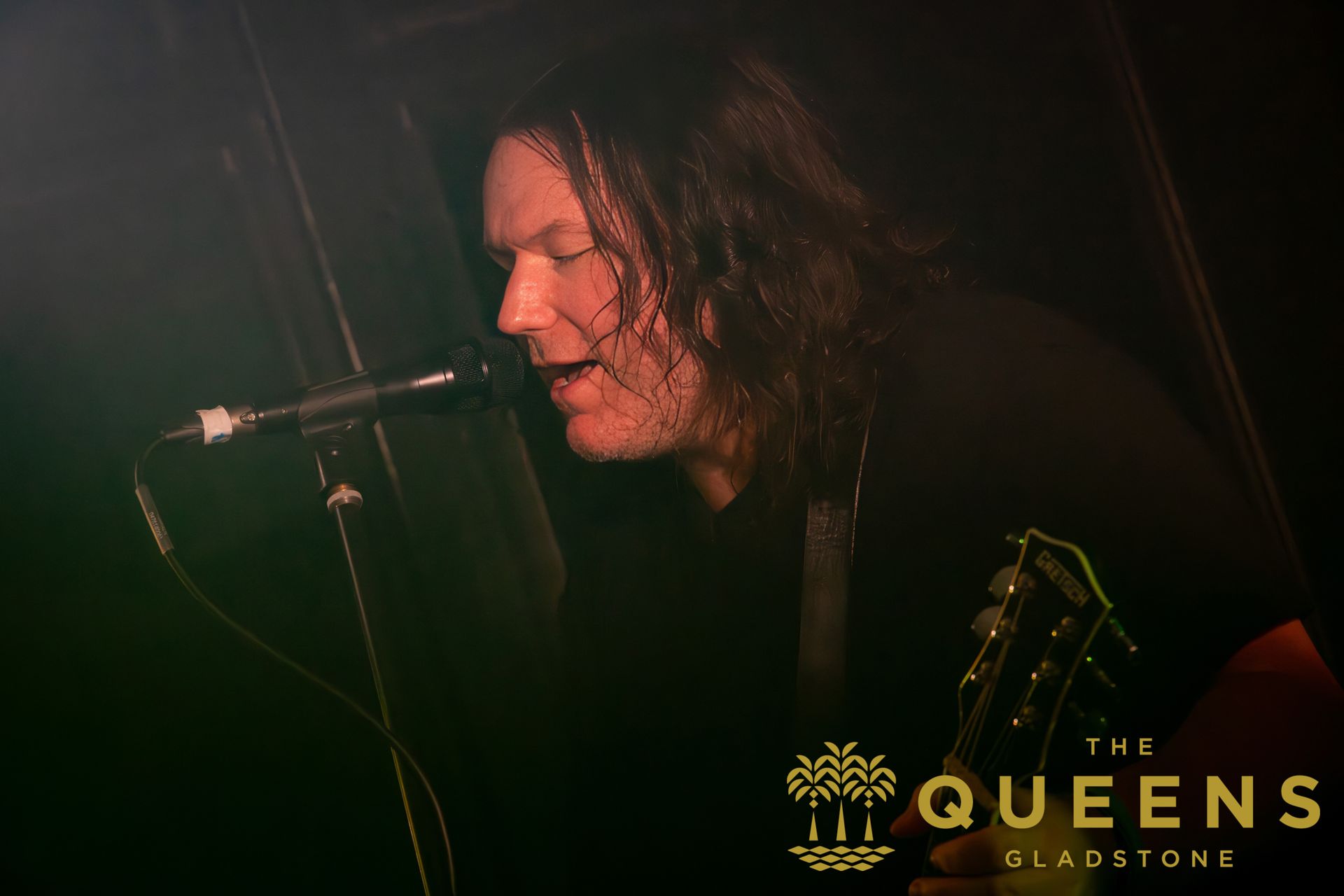 Singer at a microphone on a dimly lit stage, with The Queens logo in the corner.