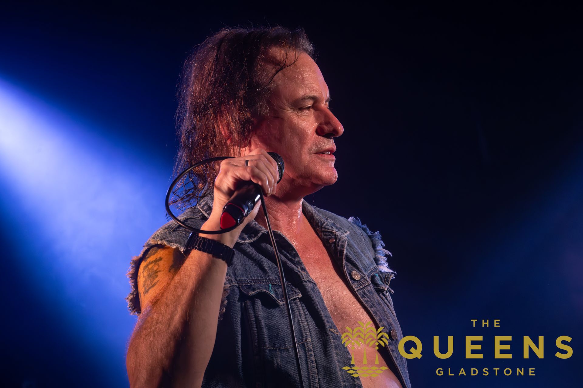 Singer performing on stage with microphone, wearing denim vest under blue and purple lights