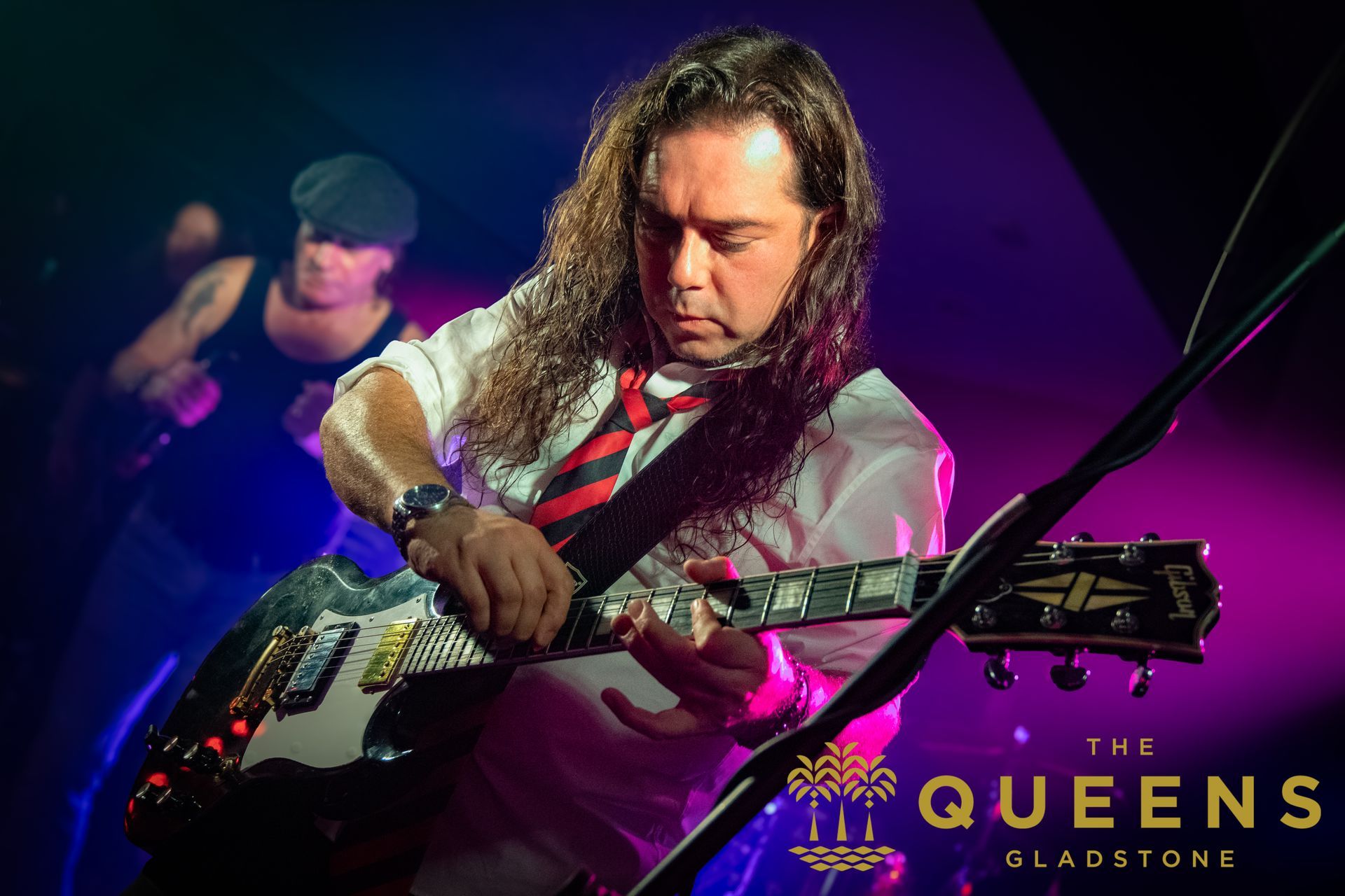 Guitarist performing on stage under purple lights, with “The Queens” sign in the background.