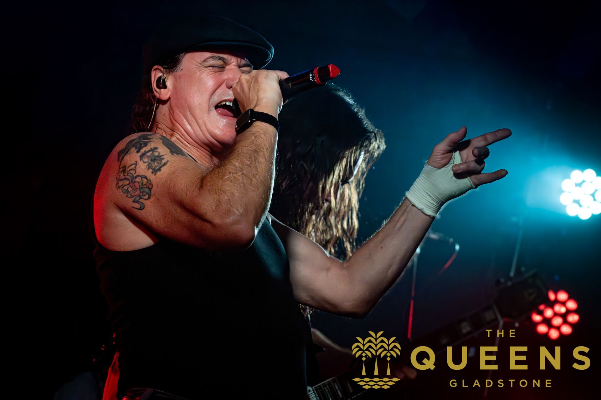 Singer performing onstage with microphone, tattoos visible, blue and red concert lights, The Queen logo in background
