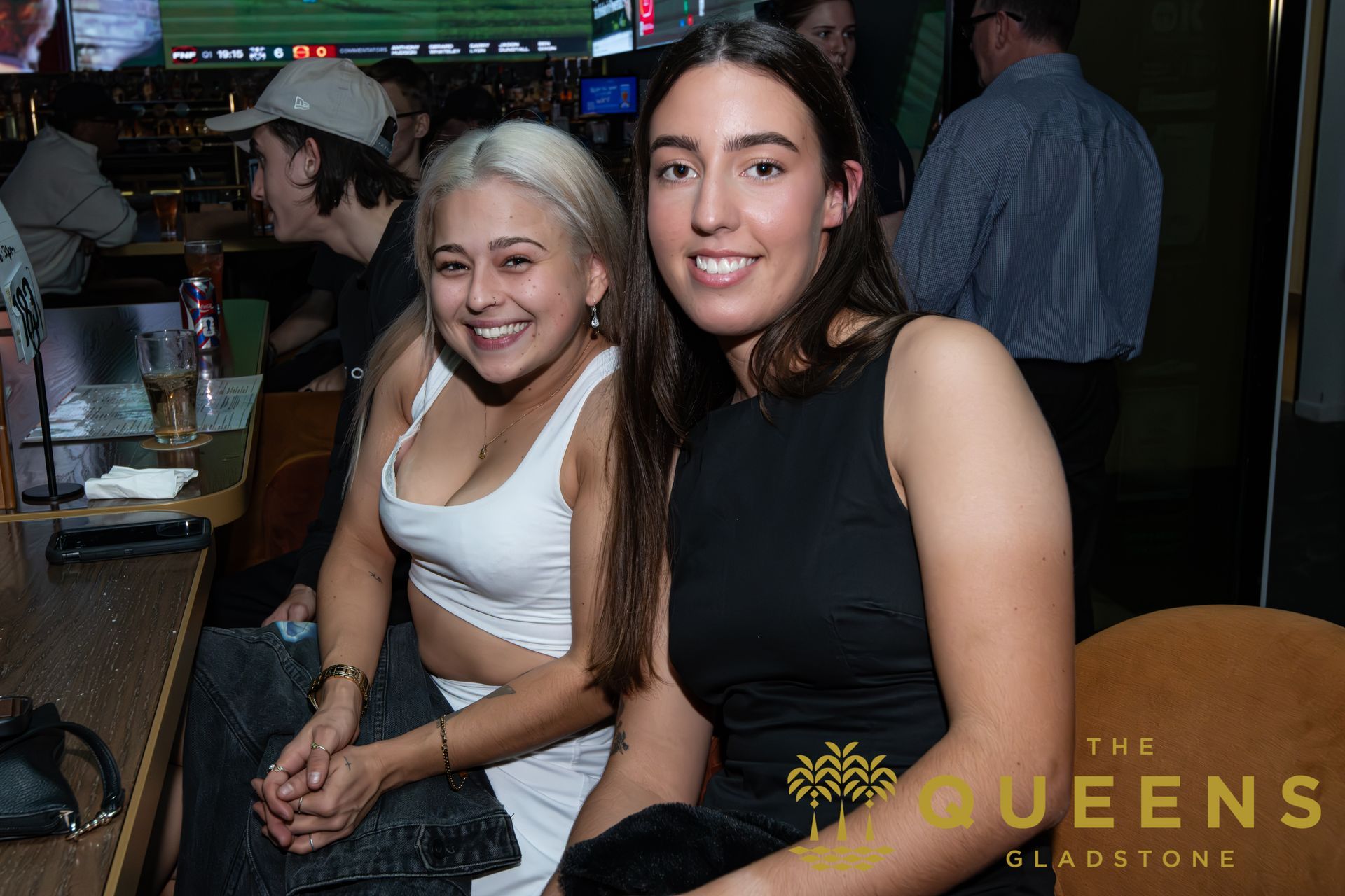 Two smiling people seated at a bar in a dim nightclub, one in white and one in black.