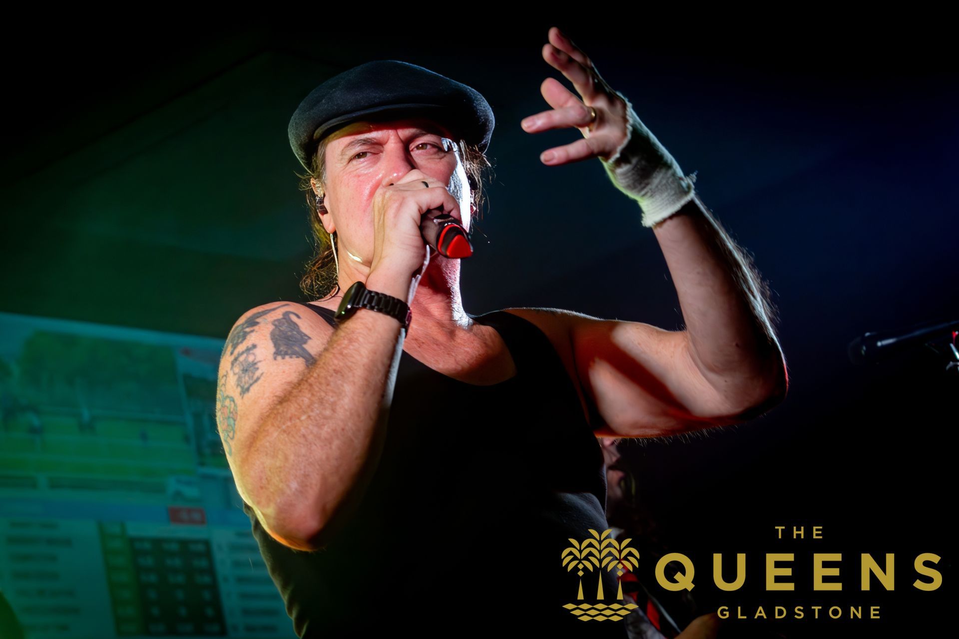 Singer performing on stage with microphone, wearing a black cap and tank top, under colorful concert lights.