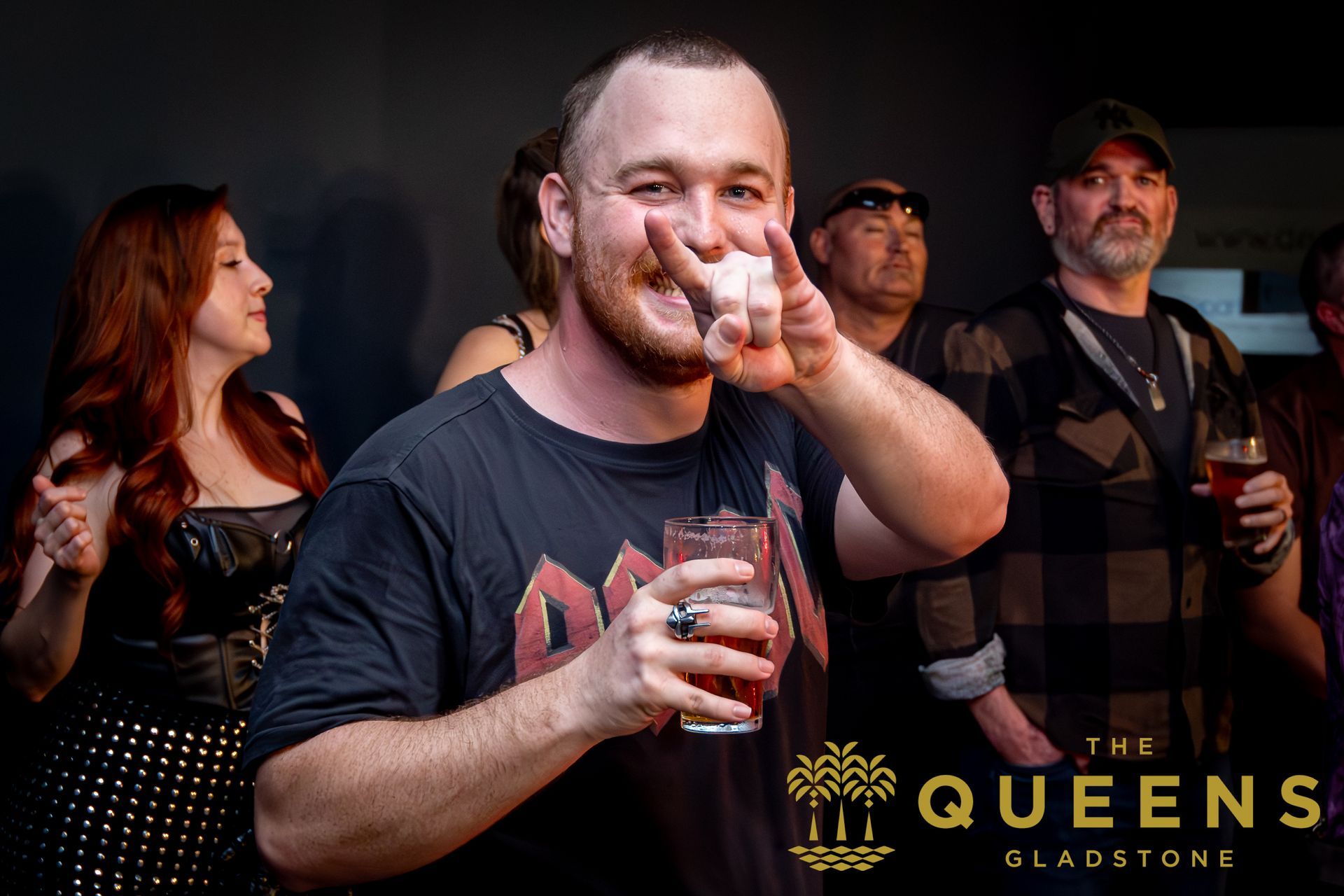 Man smiling with a drink and pointing at the camera in a dim bar, others standing behind him
