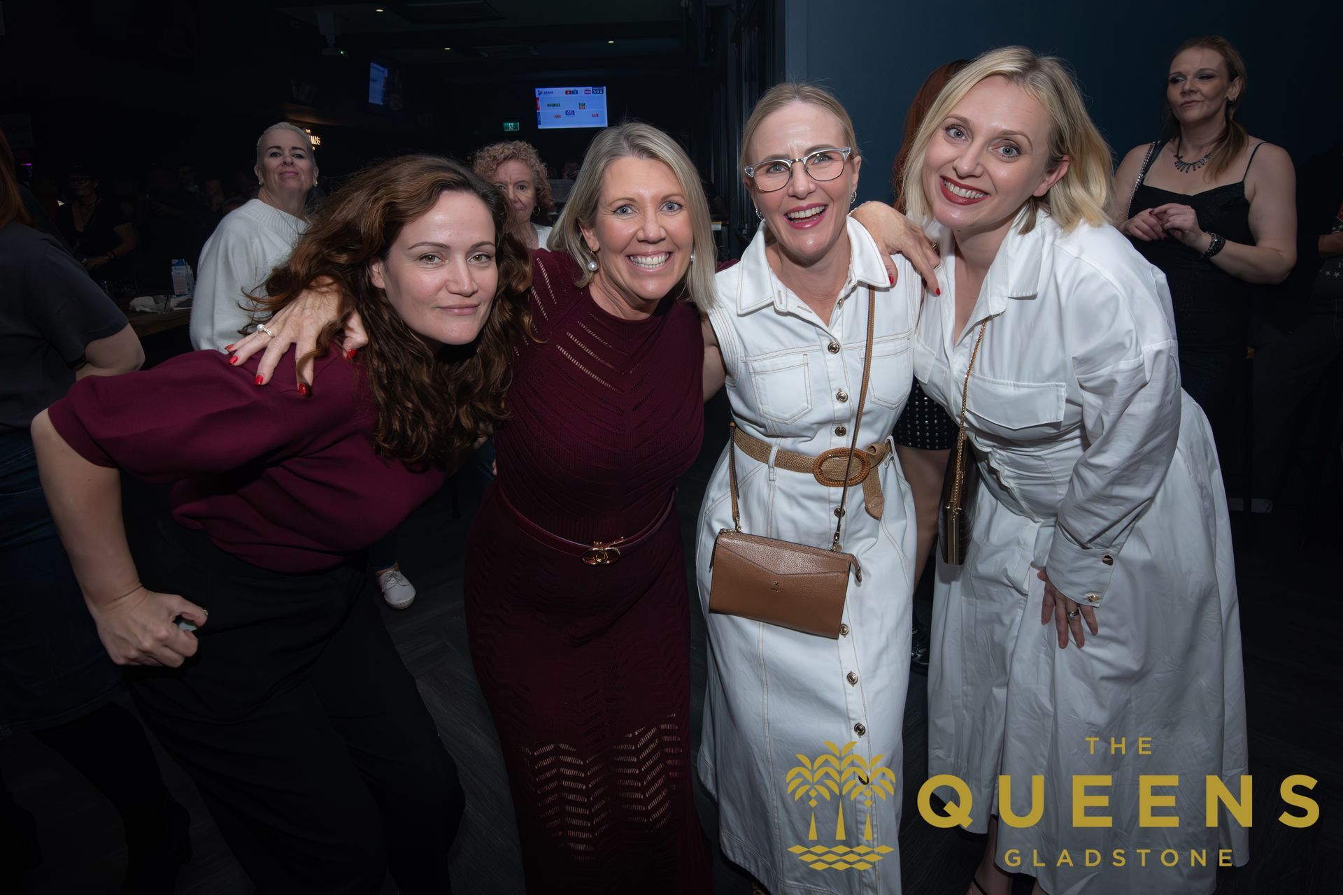 Four women posing together at a dark event, smiling in coordinated dresses and white outfits.