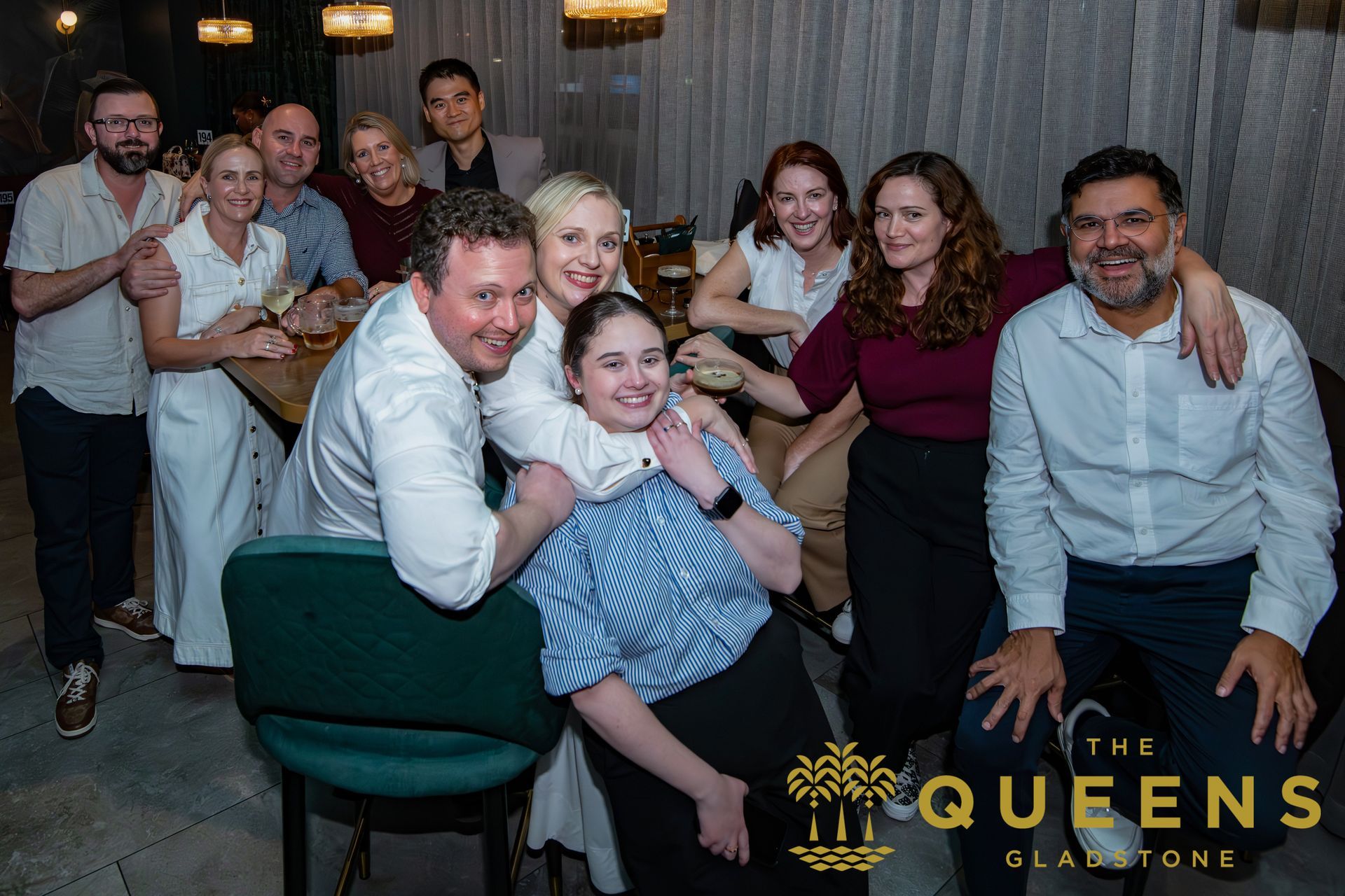 Group posing and smiling at a lively indoor event with a “The Queens” logo in the corner
