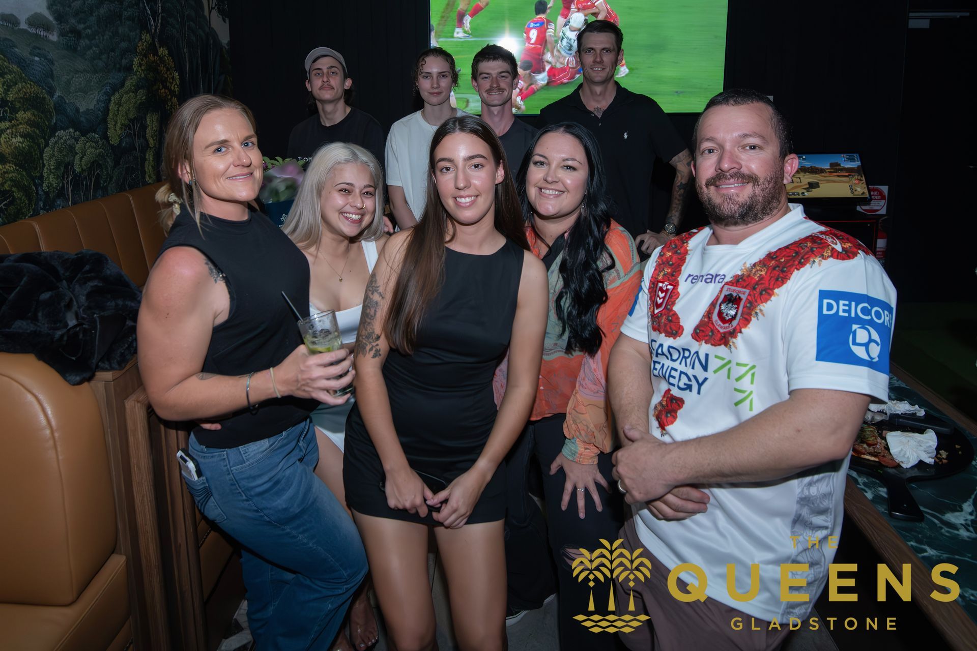 Group posing at a nightclub booth with drinks, colorful lights, and a QUEENS logo backdrop