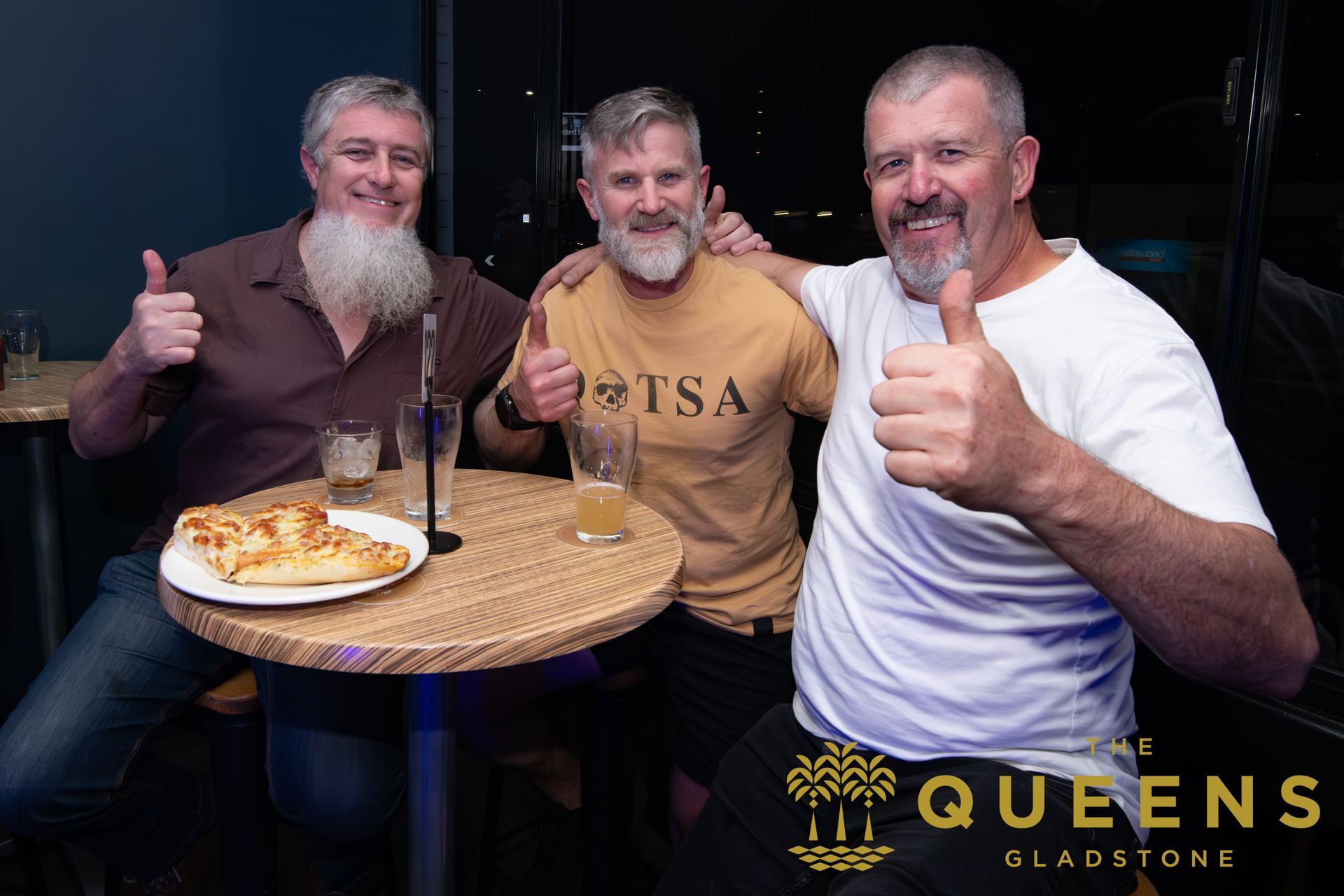 Three men smiling and giving thumbs up at a small table with food and drinks in a dimly lit venue