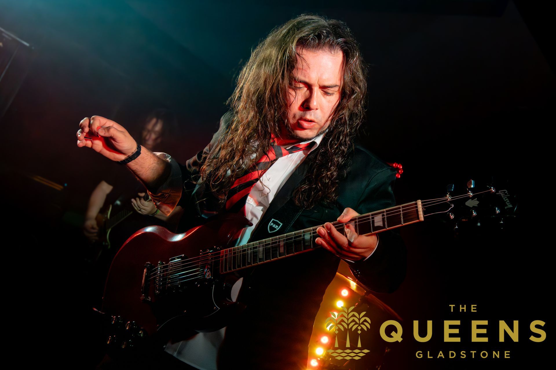 Guitarist performing under red stage lights, with “The Queens of Slapstock” logo in the corner