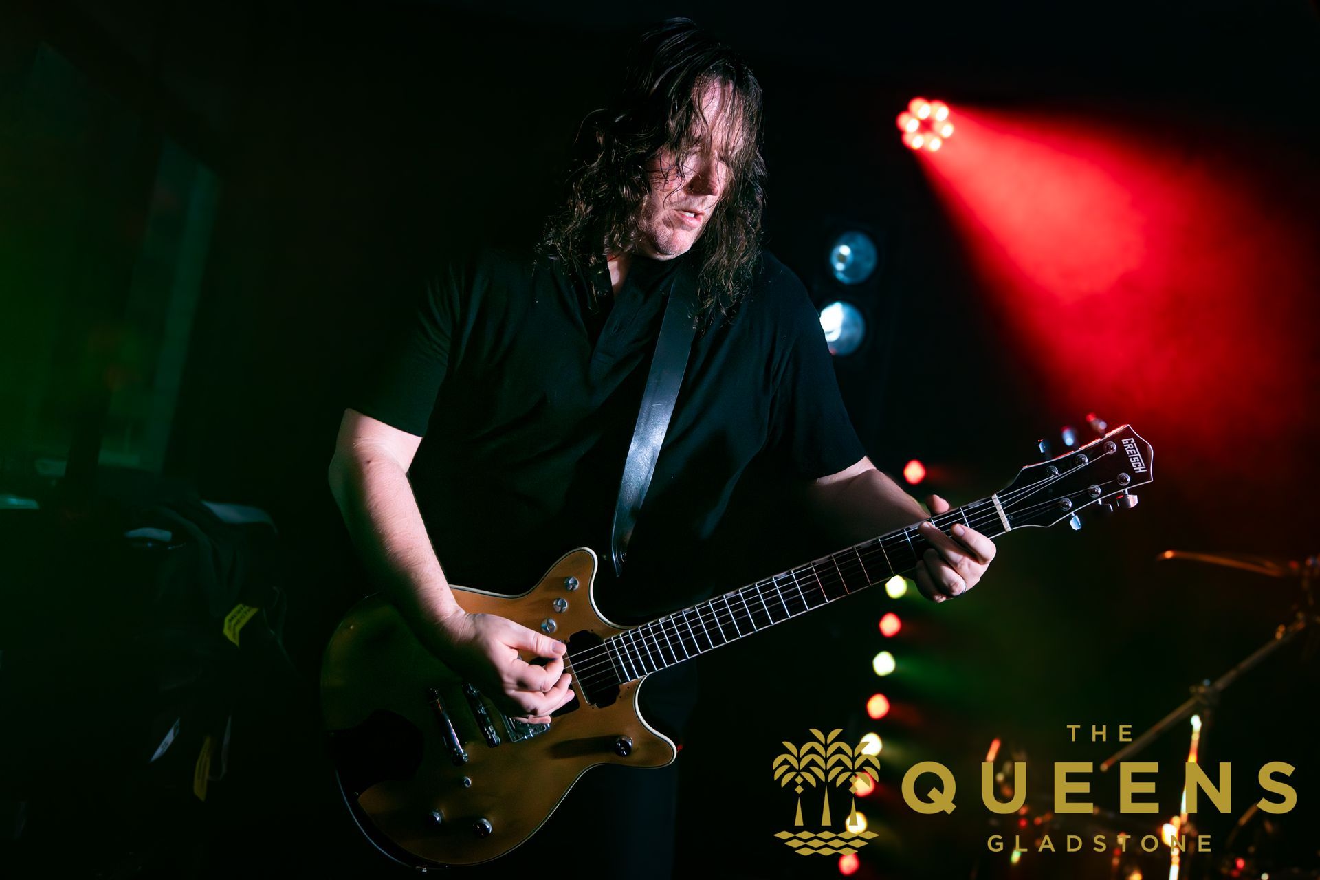 Guitarist performing onstage under red and blue lights, with “The Queens” logo in the corner