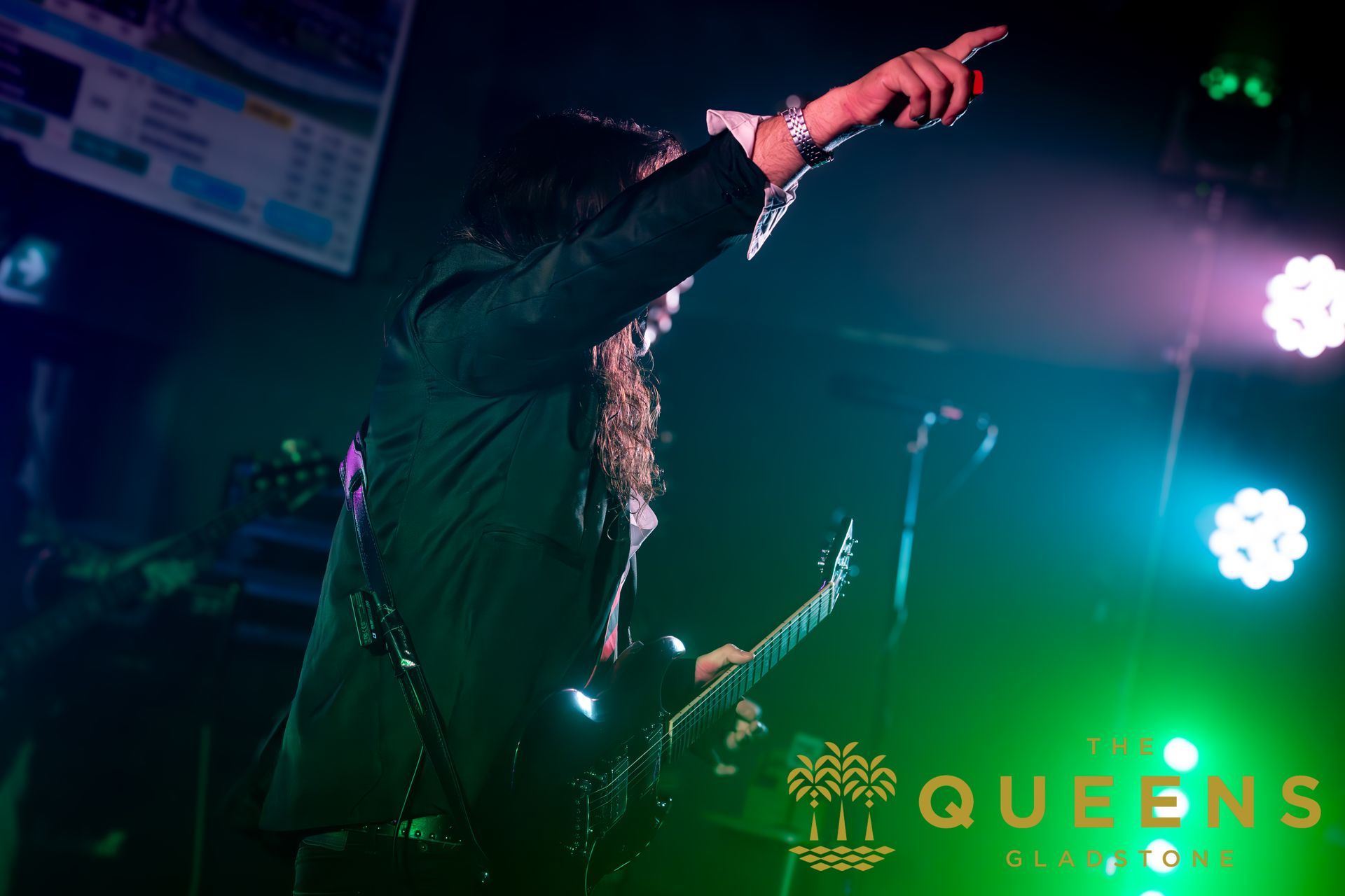 Singer onstage pointing into the crowd under pink and green lights, with “QUEENS” lit on the backdrop.