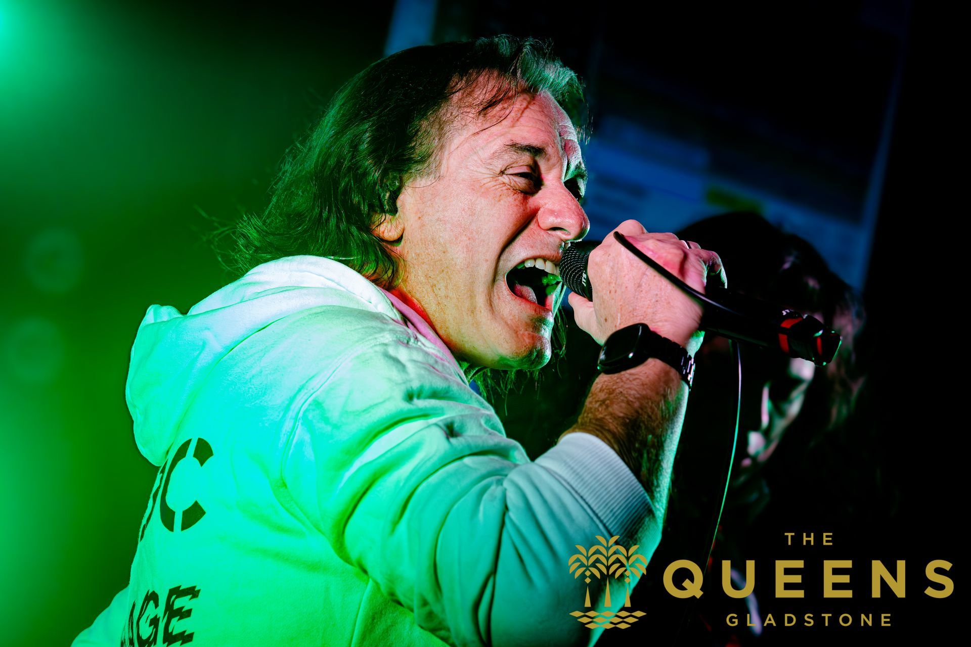 Singer performing on stage under green lights, holding a microphone with “The Queens” logo in the corner
