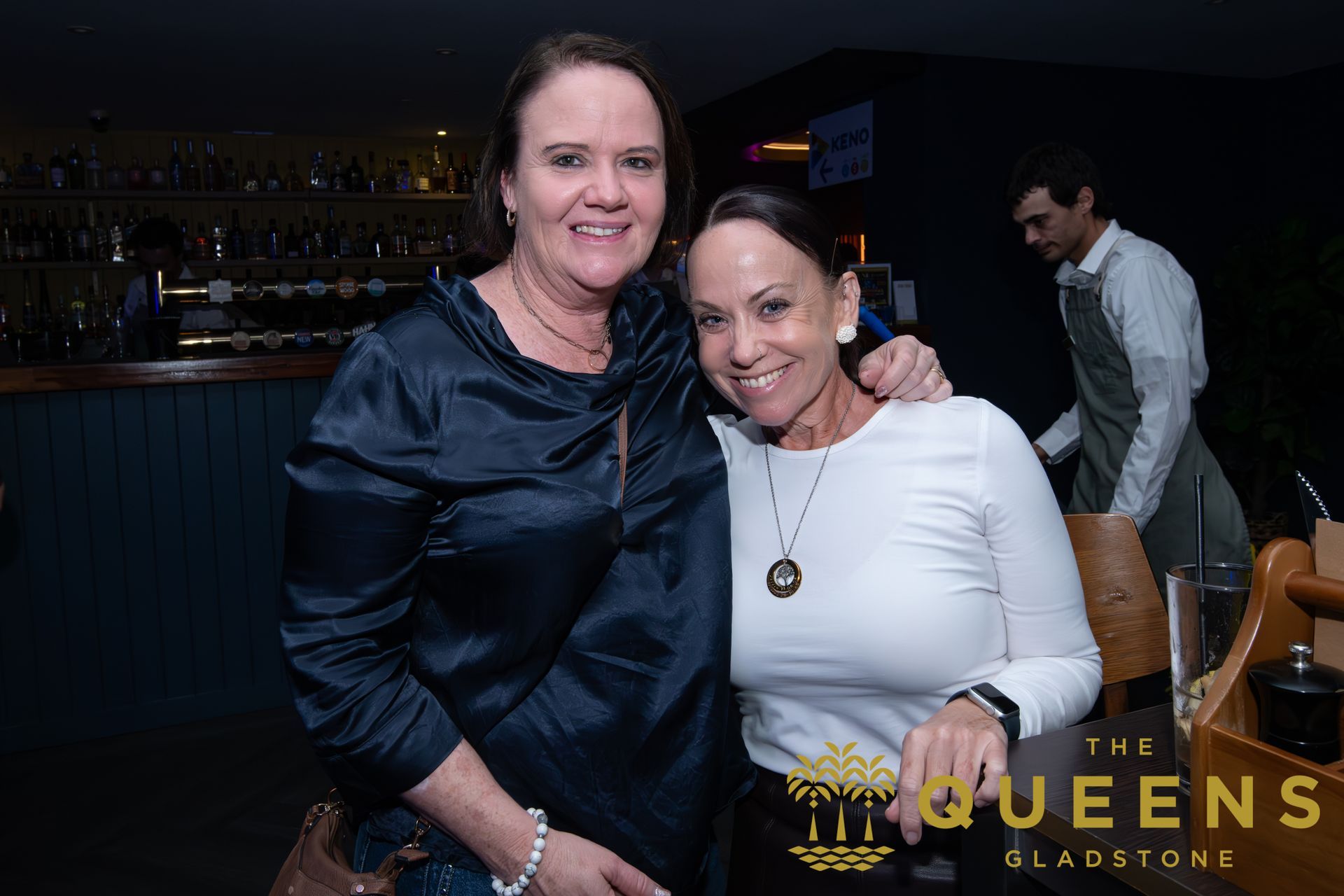 Two women posing together at a dimly lit event bar, smiling with drinks nearby.