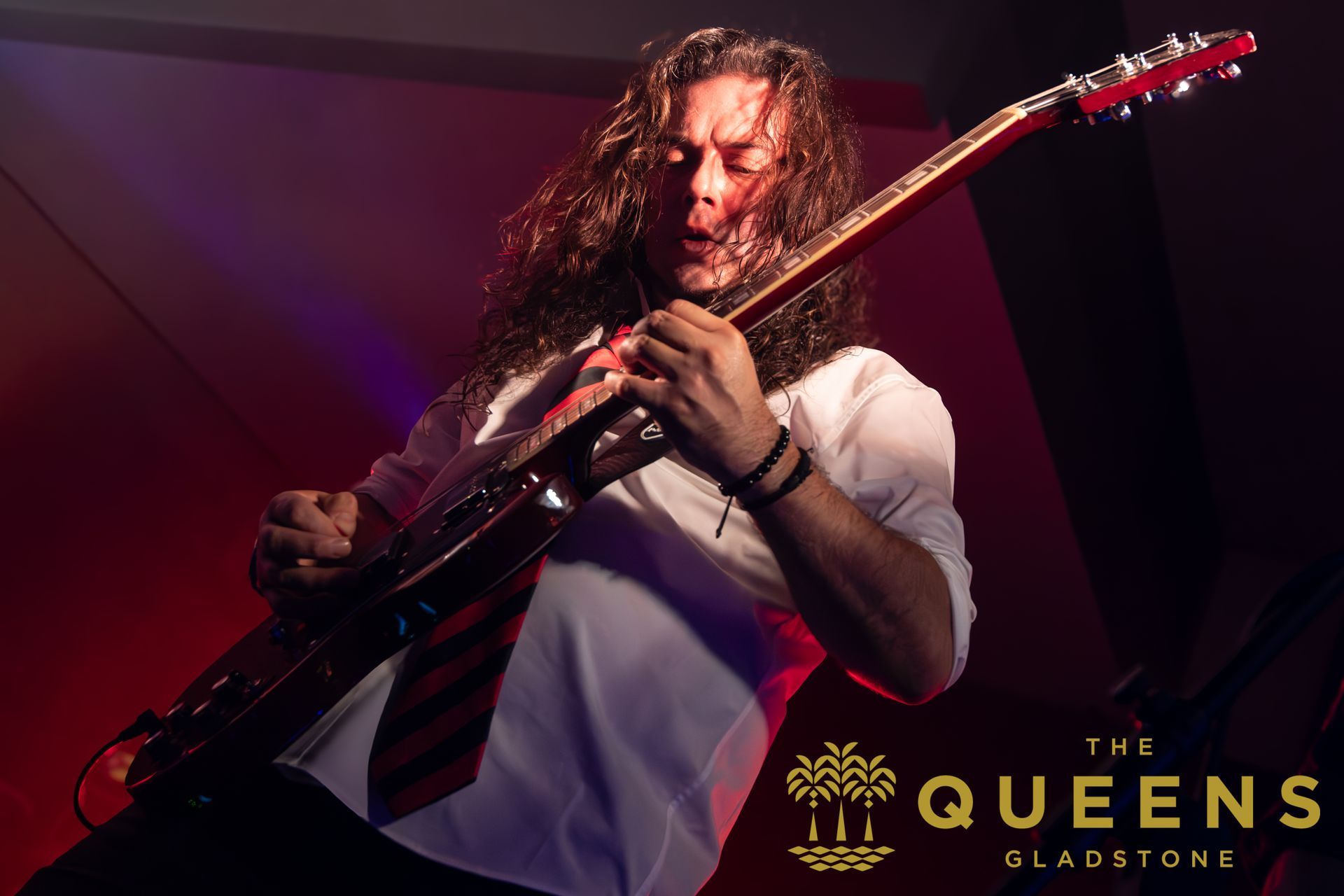 Guitarist performing under purple stage lights with “The Queens” logo in the corner