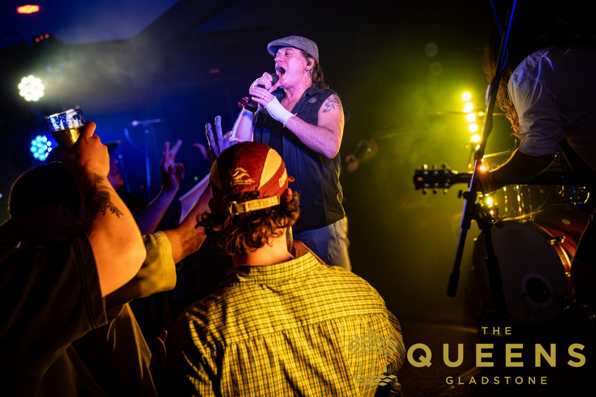 Live band performing on a dimly lit stage with purple and yellow lights, audience raising hands in front