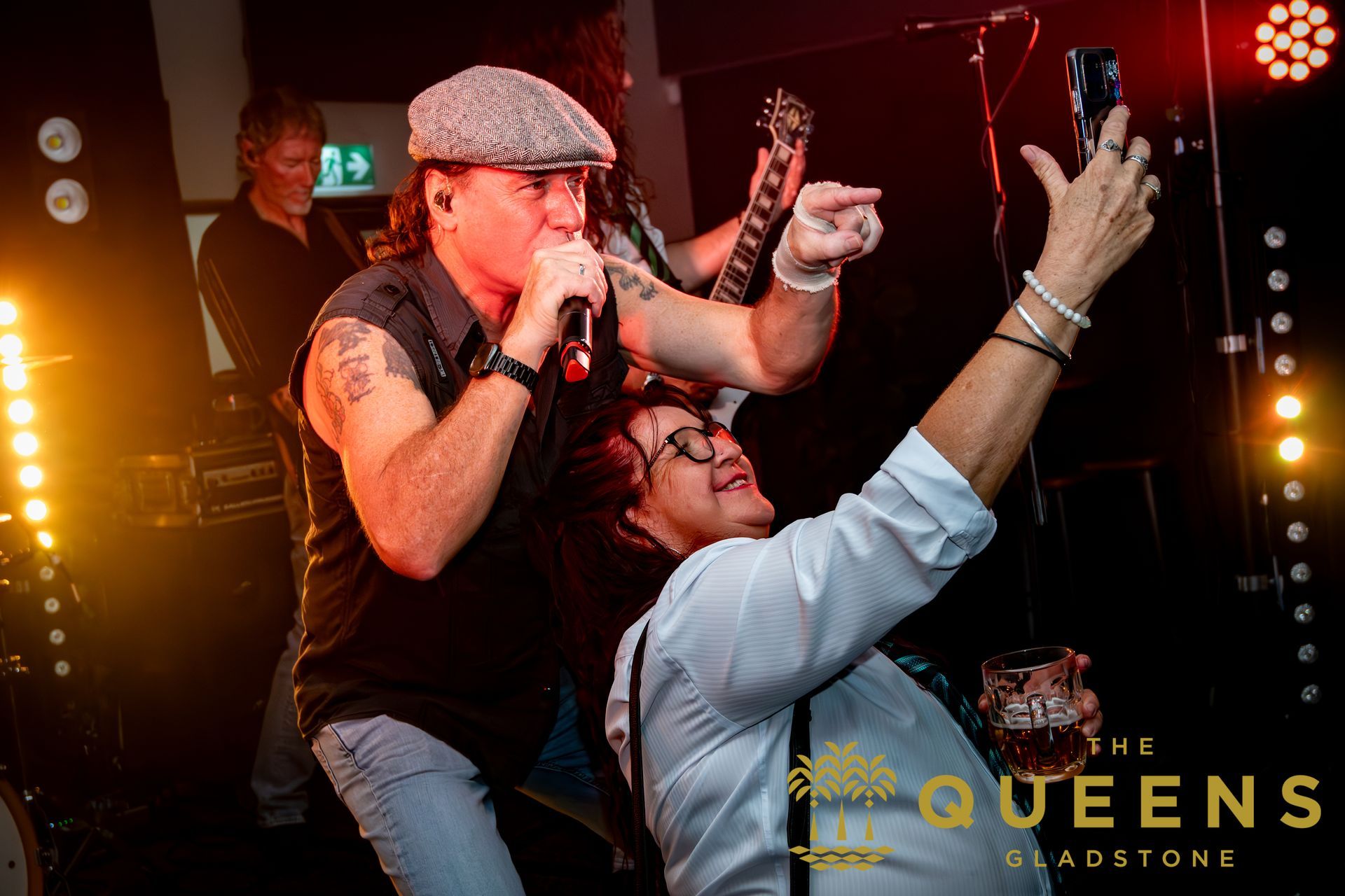 Singer performing onstage with raised arms, backed by warm stage lights and a “QUEENS” logo