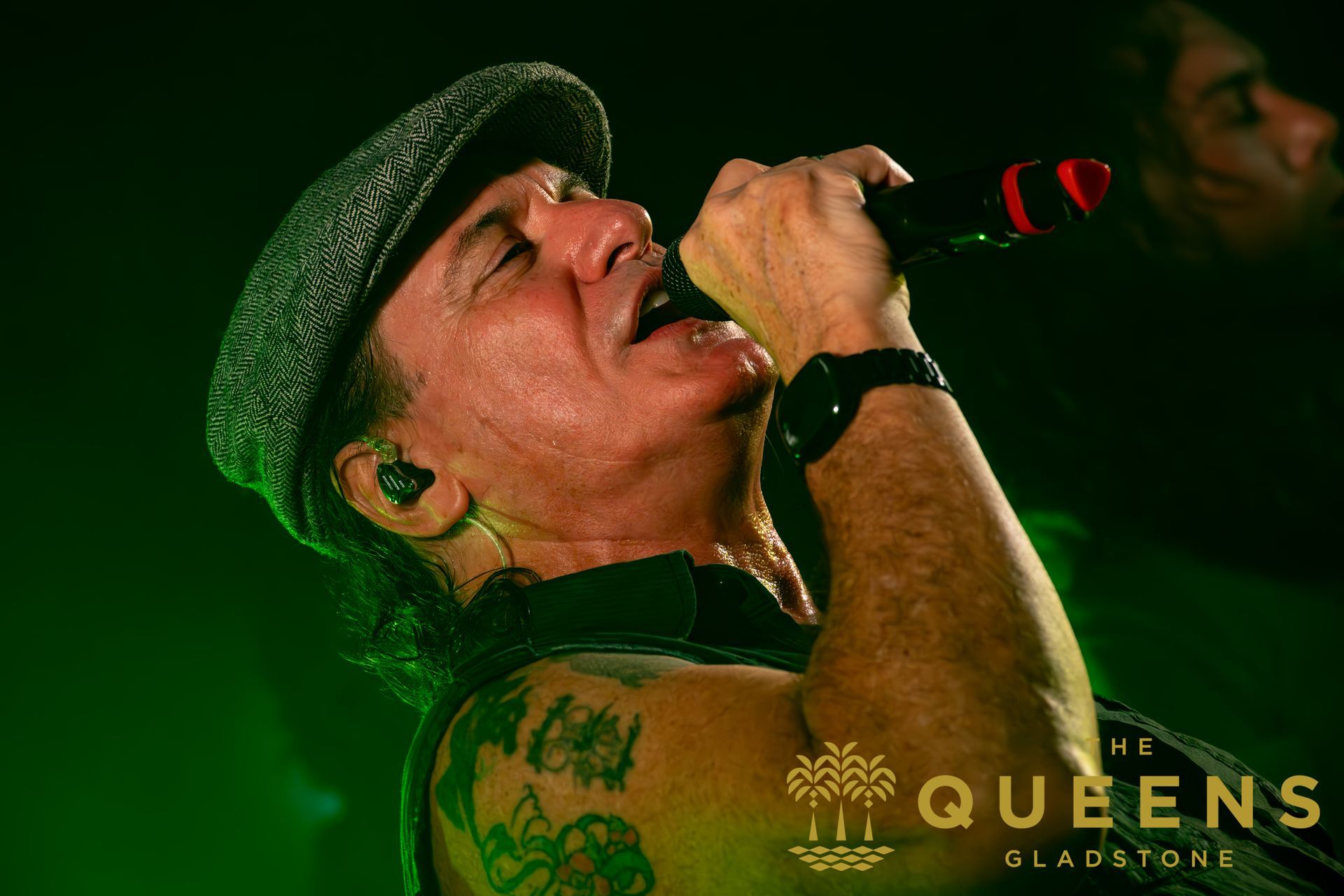 Singer in a flat cap performing into a microphone under green stage lighting, with a QUEENS logo.