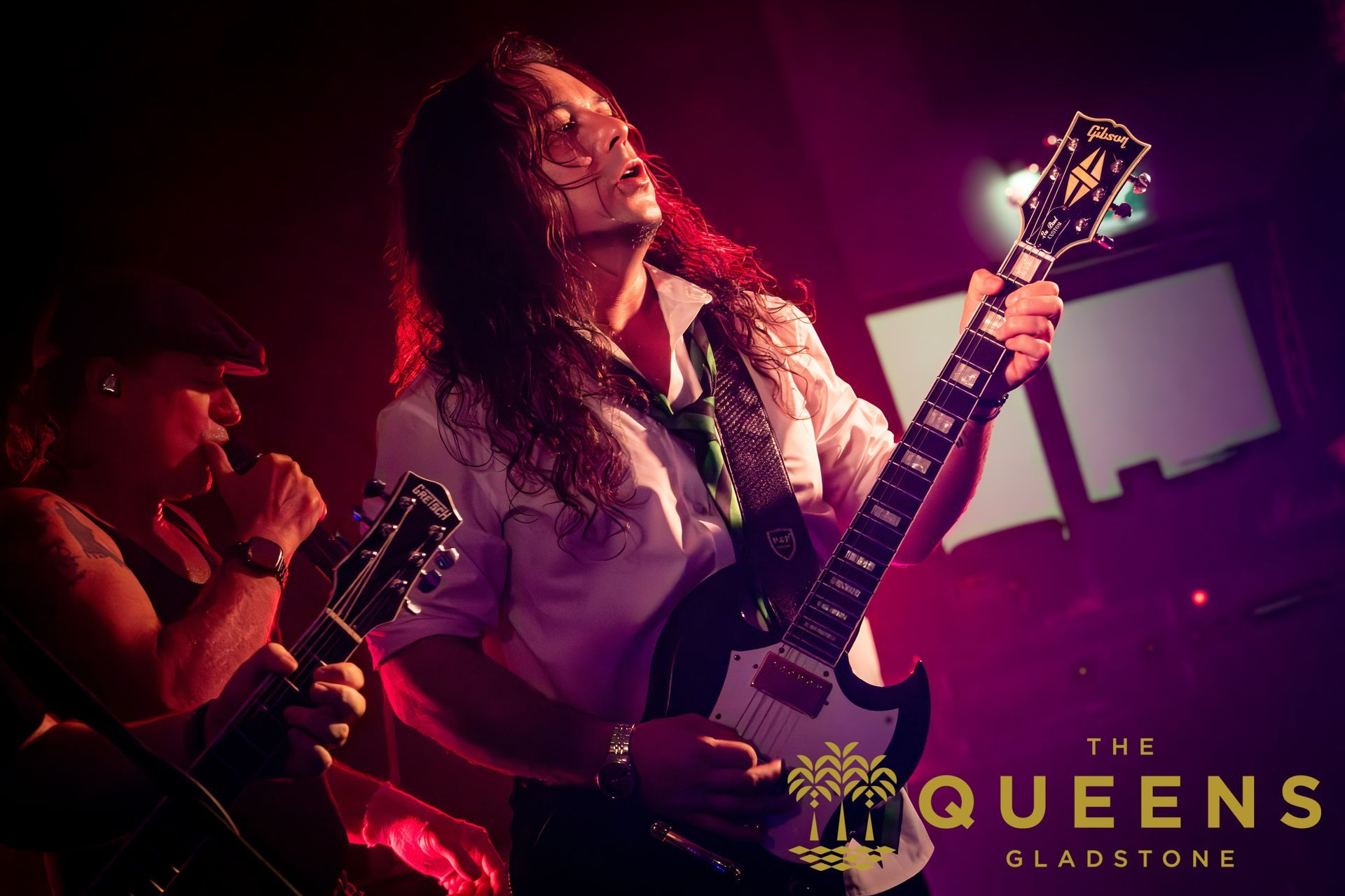 Live band performance in red stage lighting, two guitarists singing and playing with The Queens logo in the corner