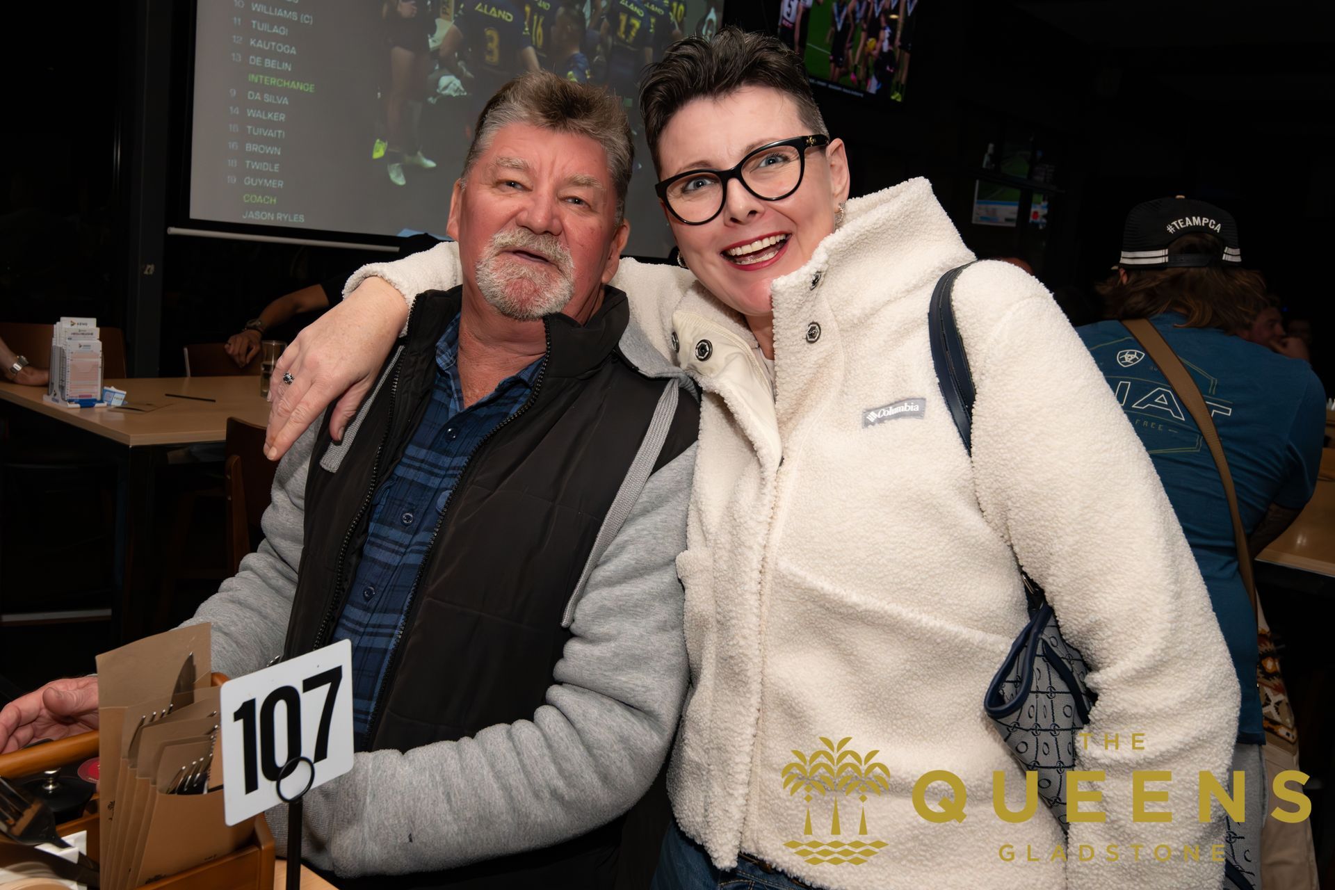 Two people smiling and posing together at an event, with a screen and “Queens” sign in the background.