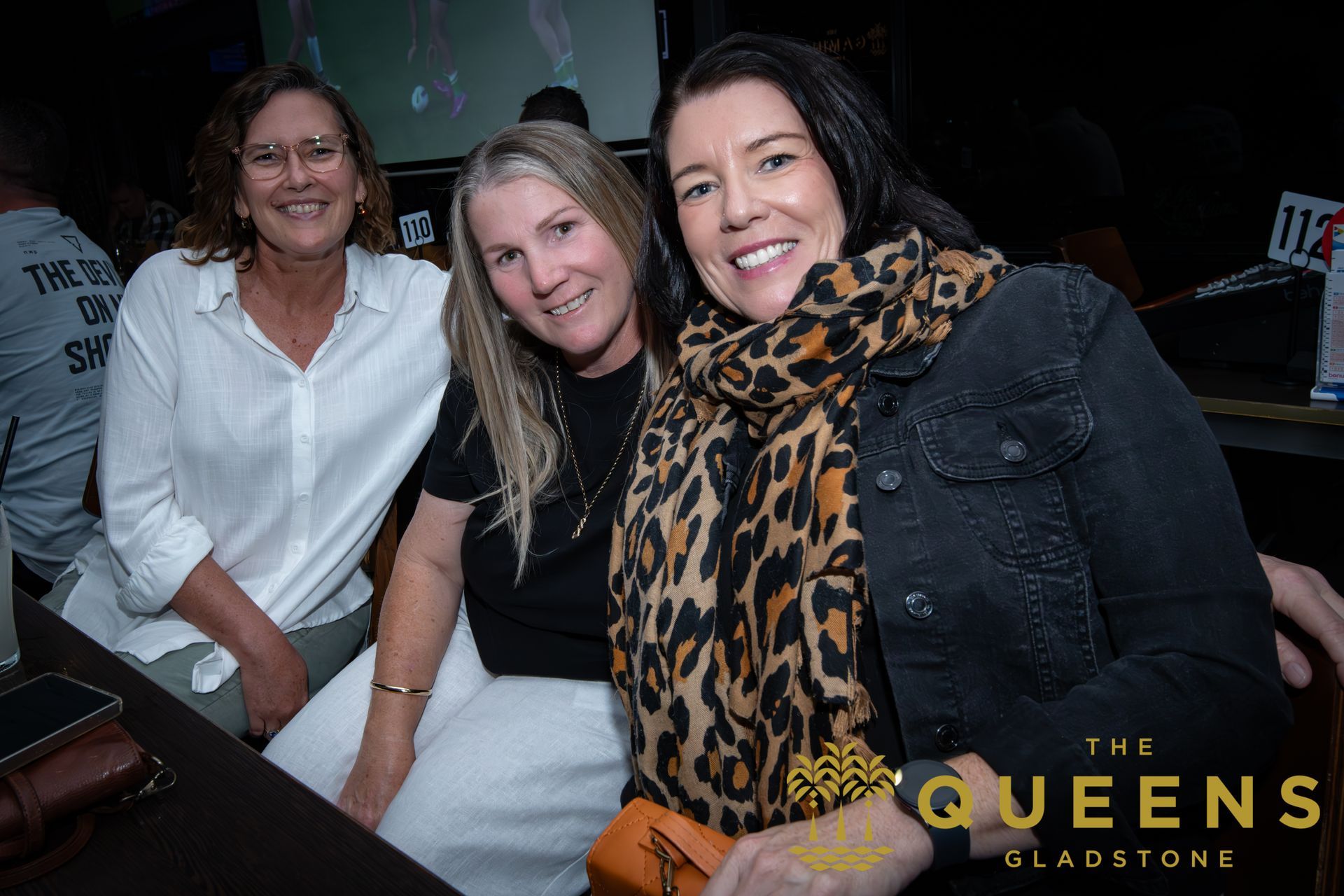 Three women smiling at an indoor event, with a gold “The Queens” logo in the corner.