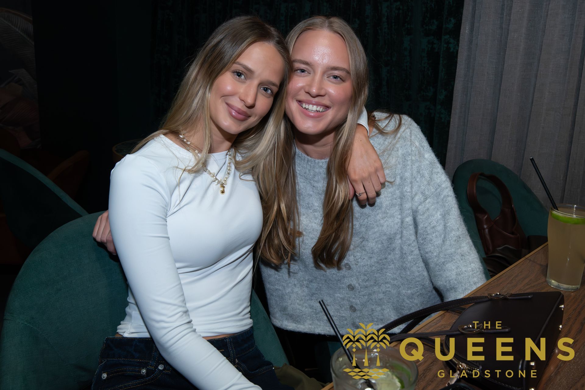 Two women smiling and posing together at a table in a dimly lit venue, with a Queens logo overlay.