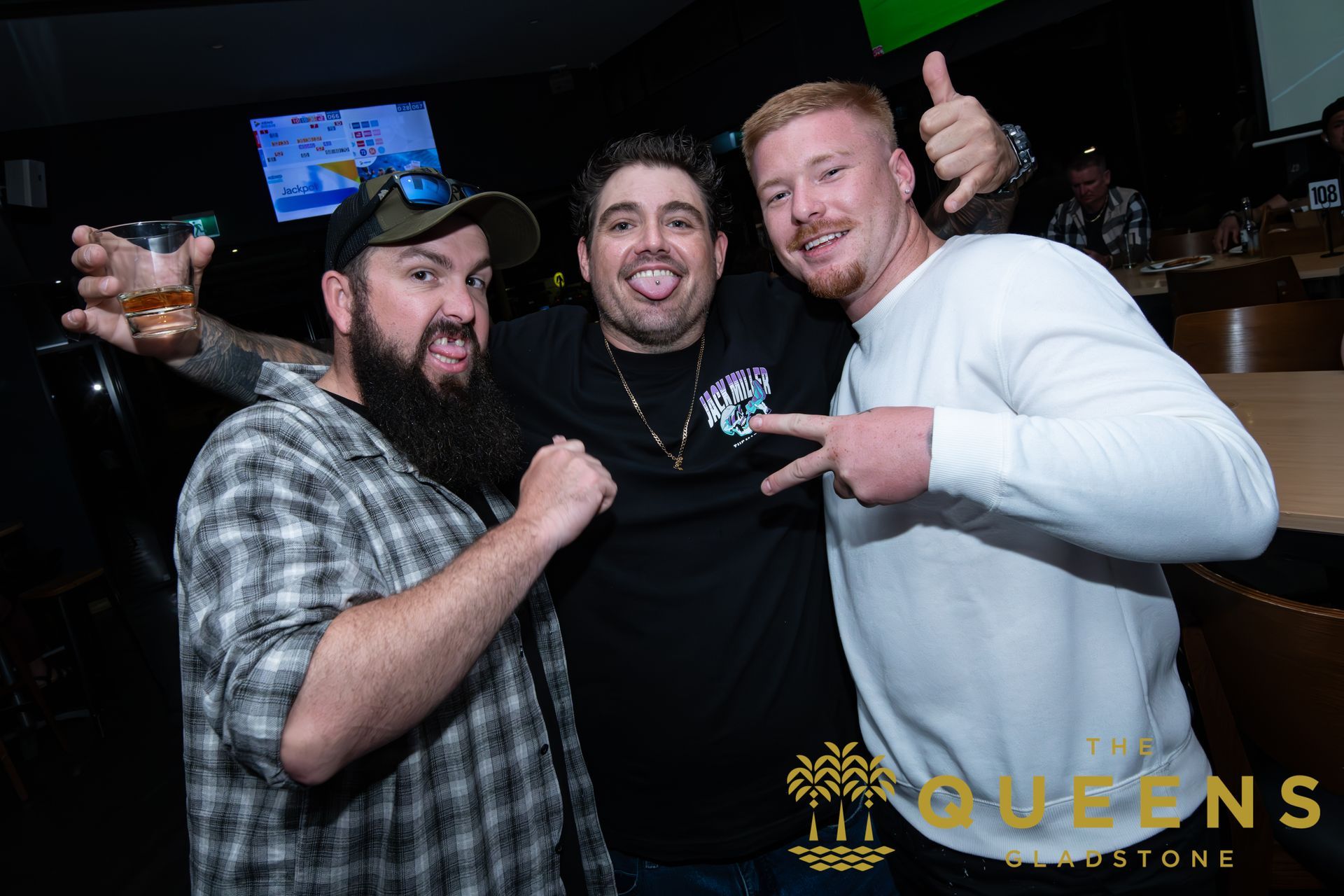 Three men posing together at a bar, smiling and pointing at each other, with drinks in hand.