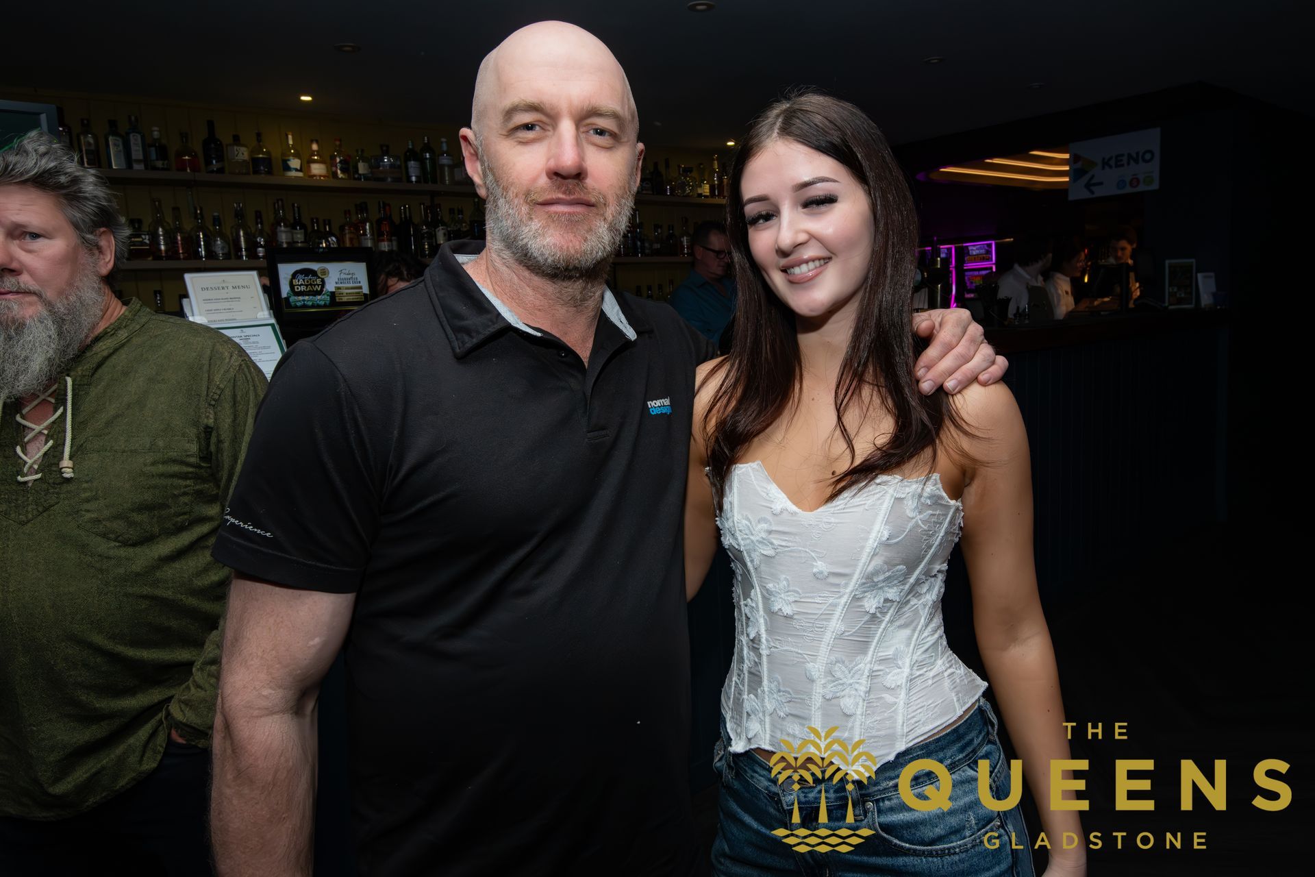 Two people posing at a bar event, with a “The Queens” logo in the bottom right.