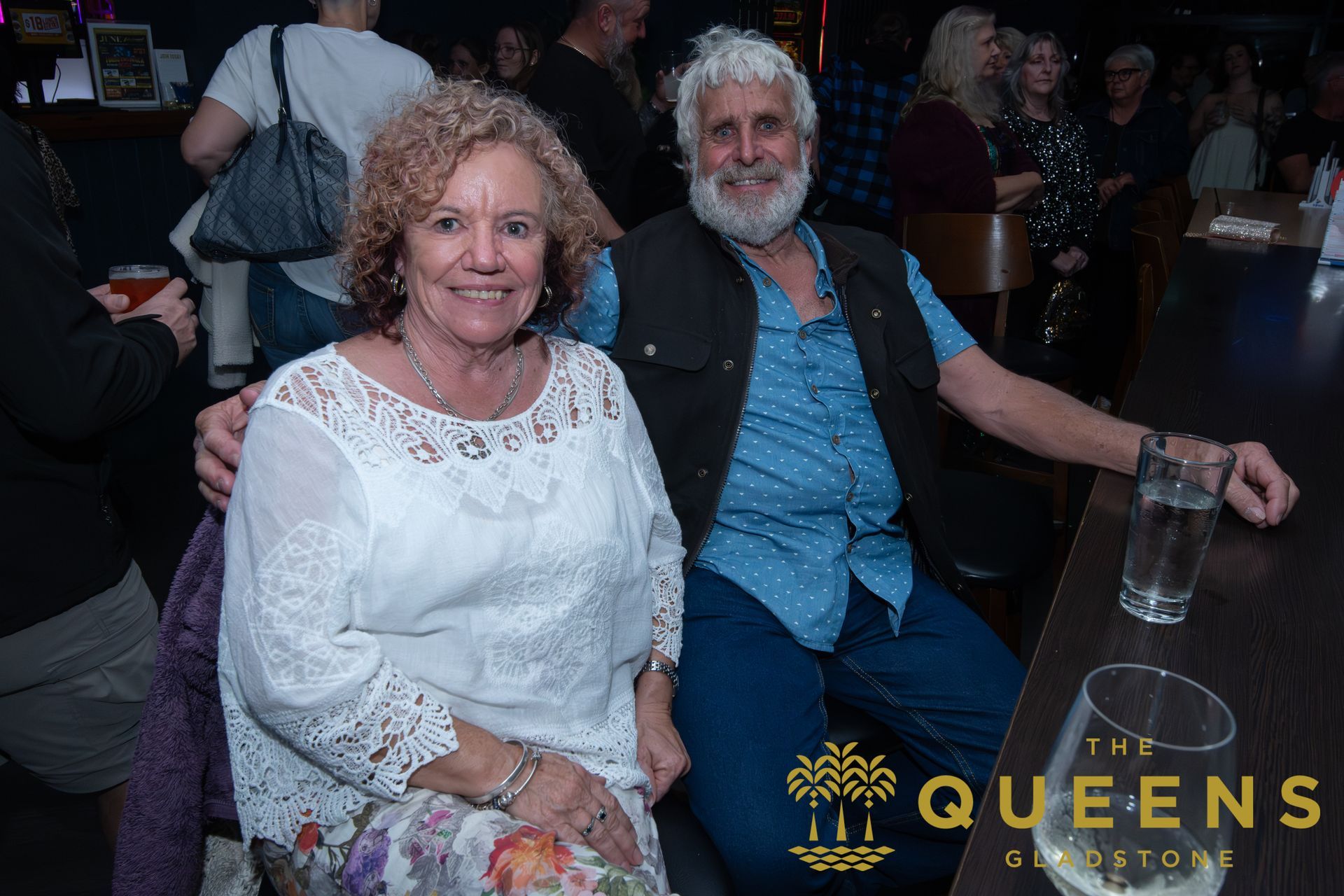 Two people seated at a dim event, smiling; “THE QUEENS” logo appears at bottom right.