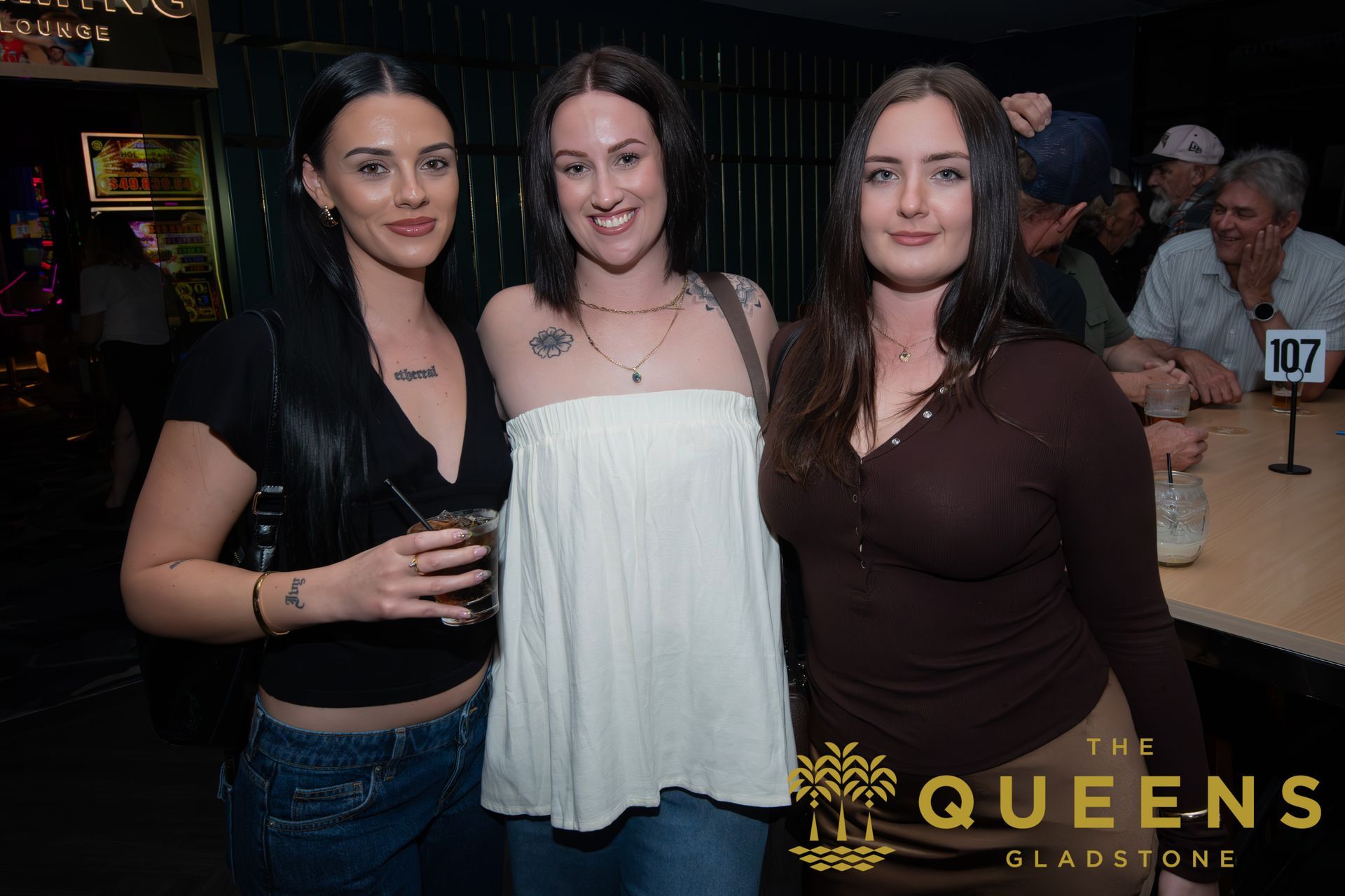Three women smiling at a nightclub table, with “The Queens” event logo in the corner.