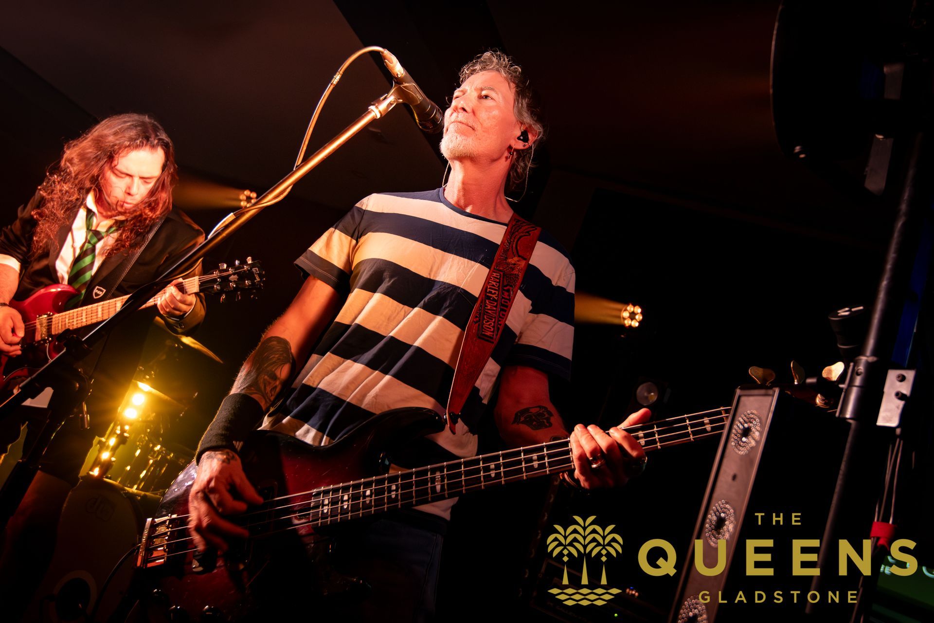 Two musicians performing on stage under warm lights, with “The Queens of…” logo at bottom right