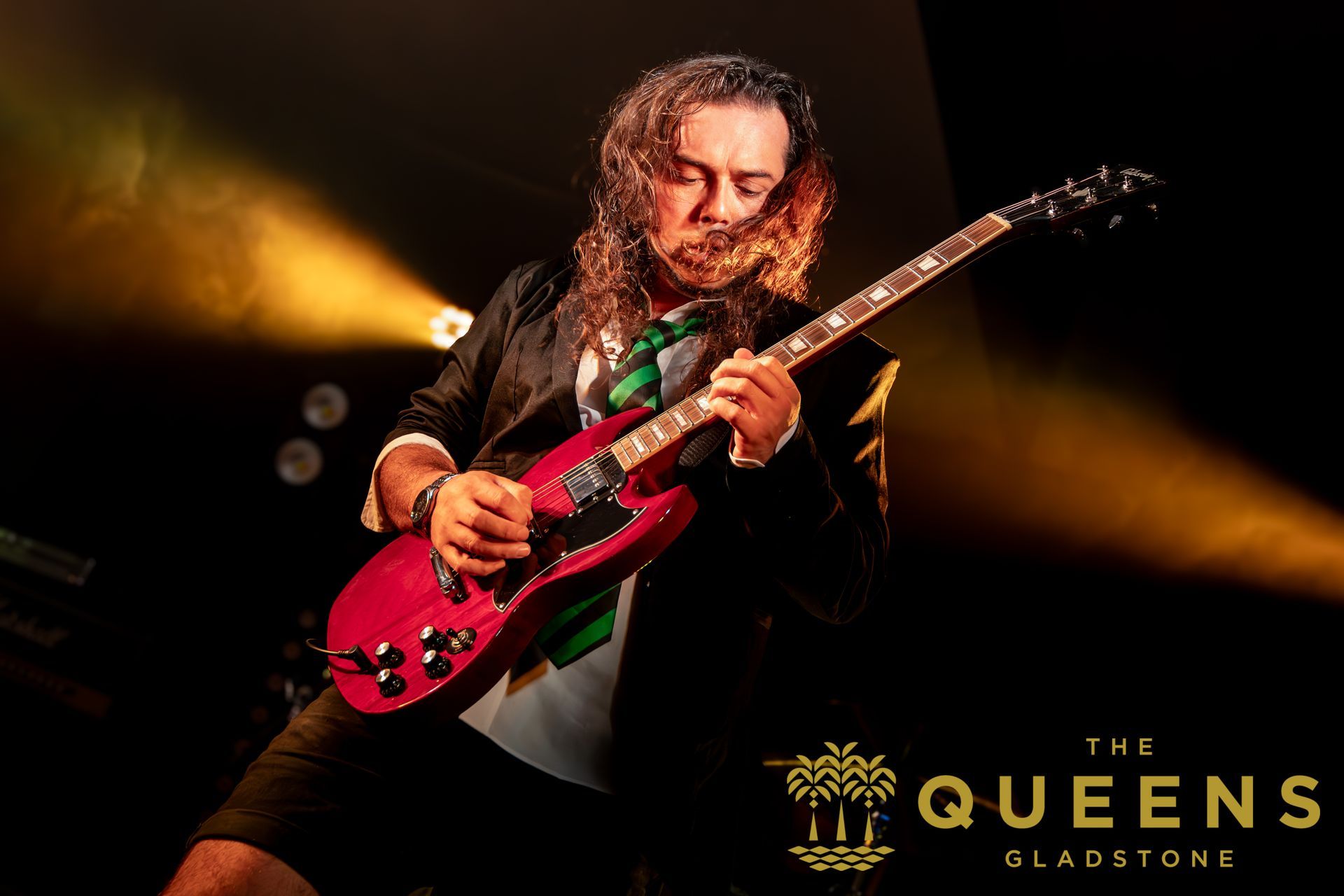 Guitarist performing onstage under warm yellow lights, playing a red electric guitar with The Queens logo