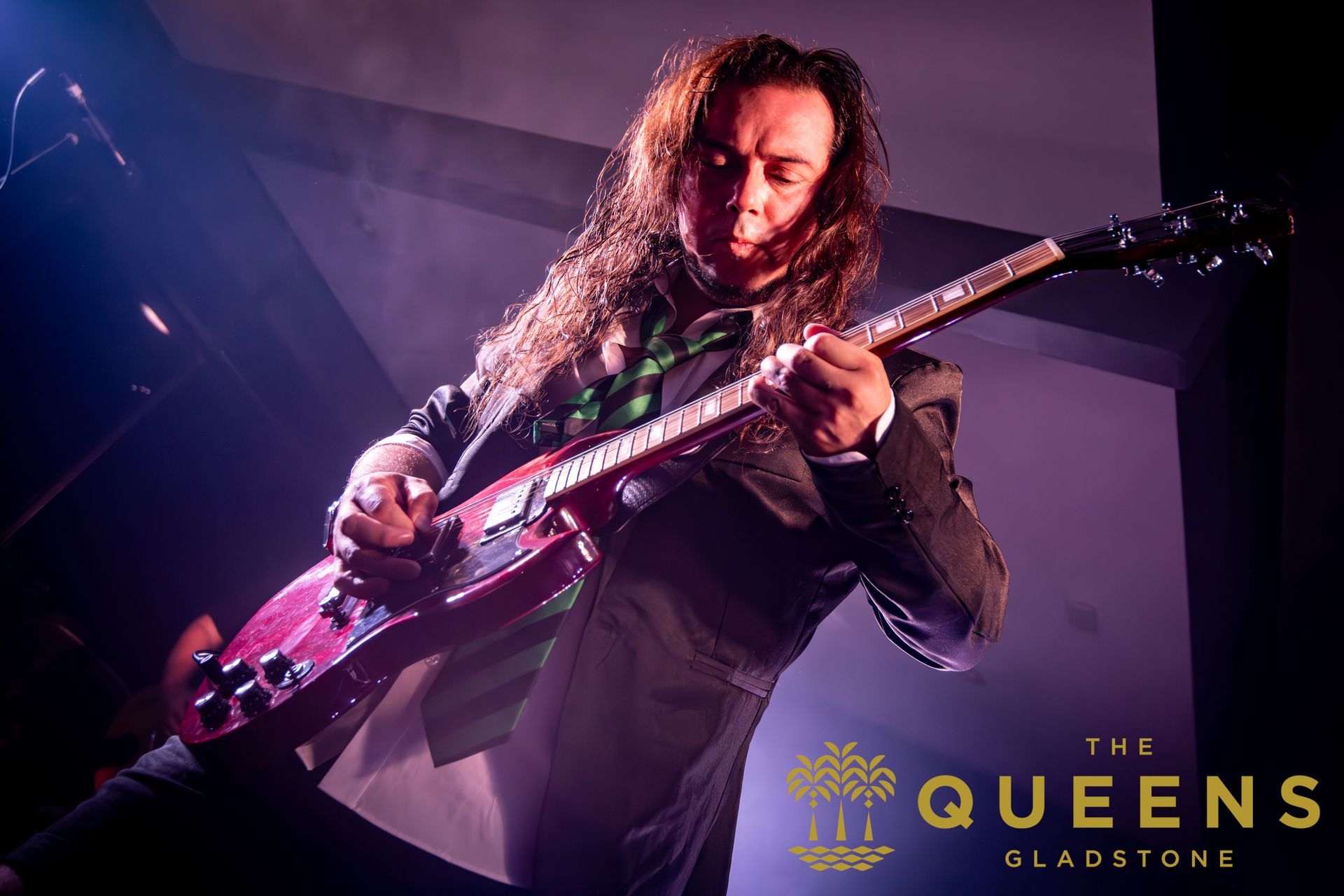 Guitarist performing on a purple-lit stage at The Queens, Sladstone, with a black and pink electric guitar