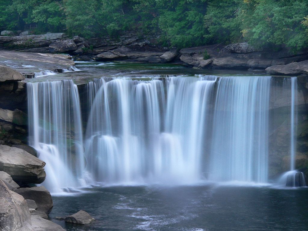 Wide waterfall cascading over a rocky ledge into a calm pool, surrounded by green trees.