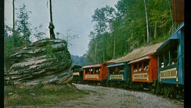 Colorful train cars on a narrow track beside a large rock and trees in a wooded valley
