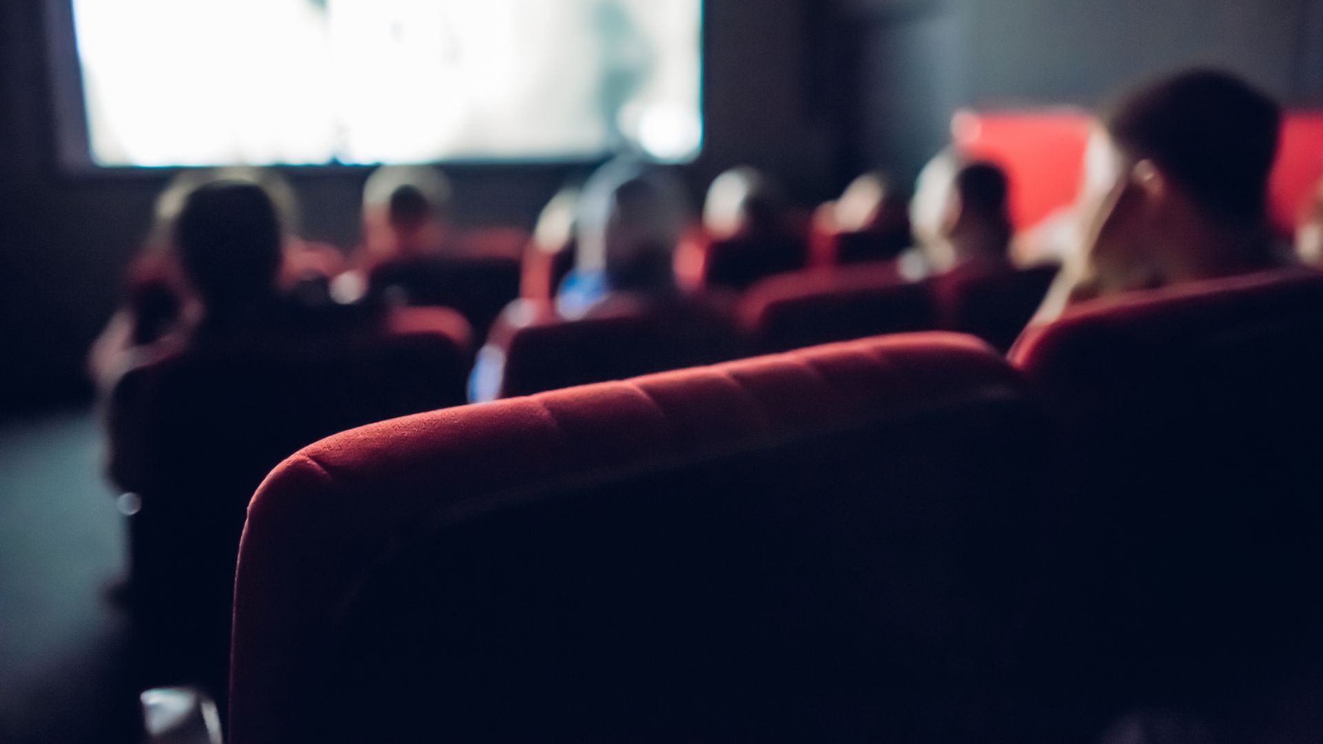 Audience seated in a dim theater, viewed from behind red seats.
