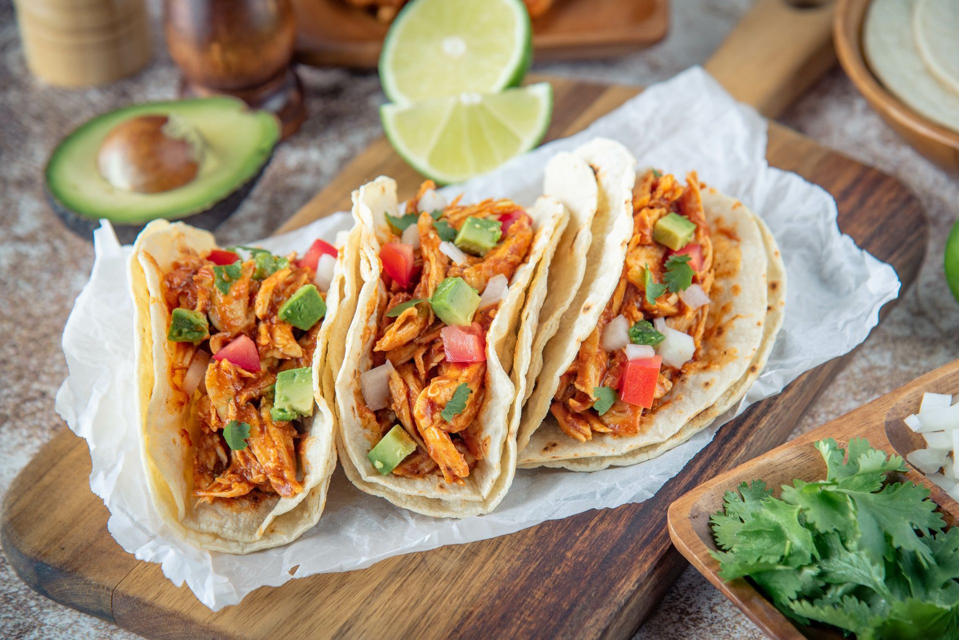 Three fish tacos on parchment, topped with salsa and avocado on a wooden board with lime wedges.