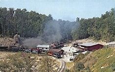 Smoke rises over a rural road with emergency vehicles and damaged buildings in a wooded area.