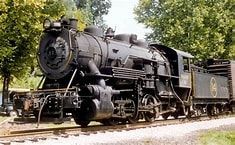 Black steam locomotive with tender on tracks, surrounded by green trees outdoors