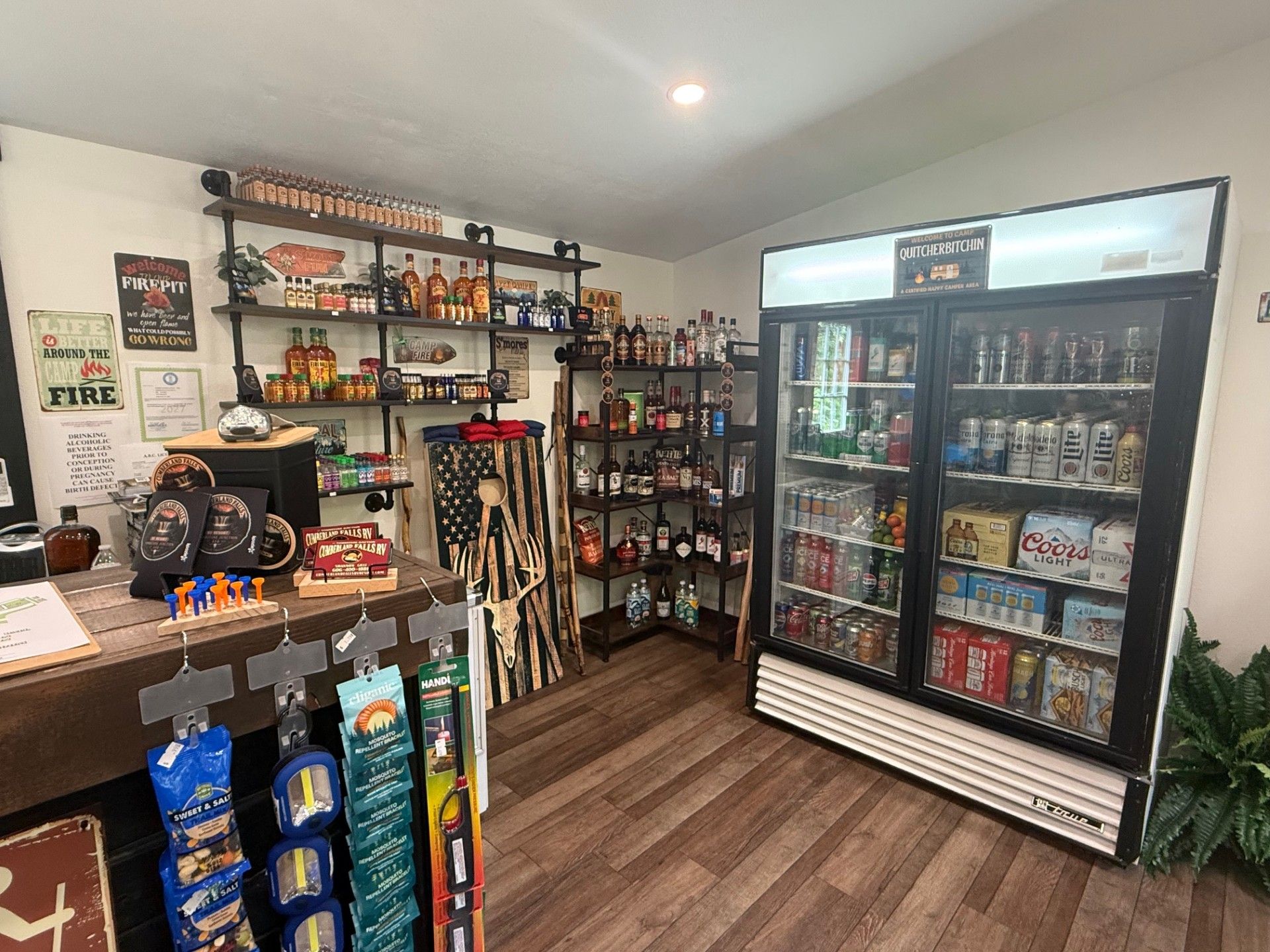Small convenience store interior with snack shelves, drink cooler, and wood-look floor.