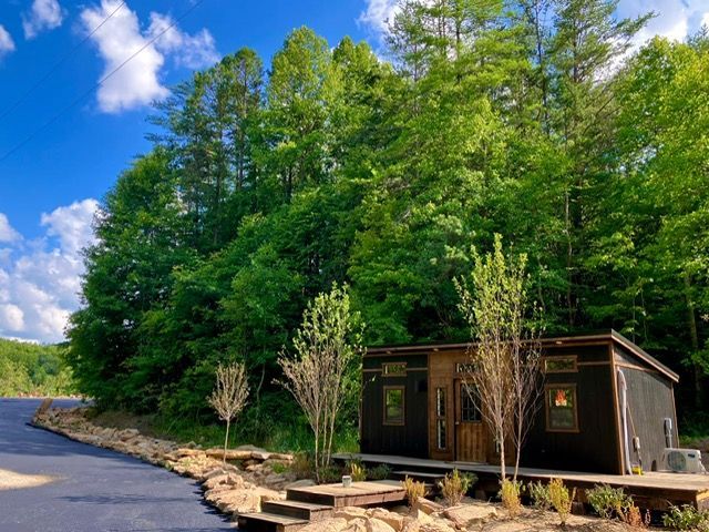 A small cabin with a wooden door sits beside a paved road and lush forest under a blue sky.
