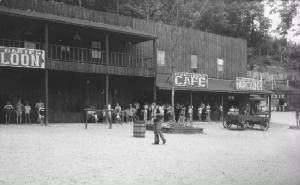 Black-and-white street scene with a saloon and cafe, people standing outside, and a car parked nearby.